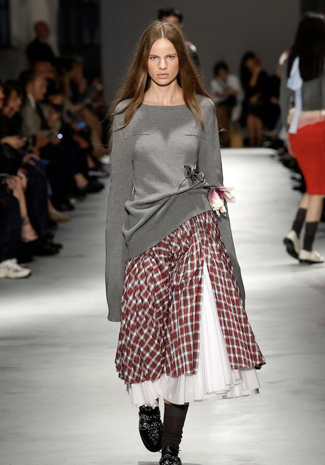 Modelo desfilando con un suéter gris holgado adornado con un detalle floral lateral, una falda a cuadros rojos y blancos sobre una capa interior blanca plisada, y zapatos oscuros con tachuelas.