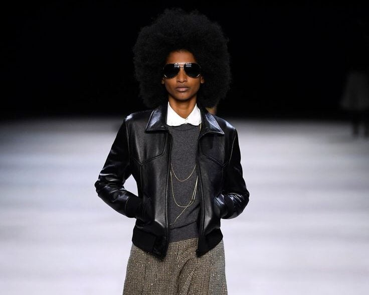 Modelo en pasarela con playera negra de manga larga desgastada, bermudas de sastre con estampado de cuadros grises, un bolso de mano negro y botines oscuros con calcetines texturizados.