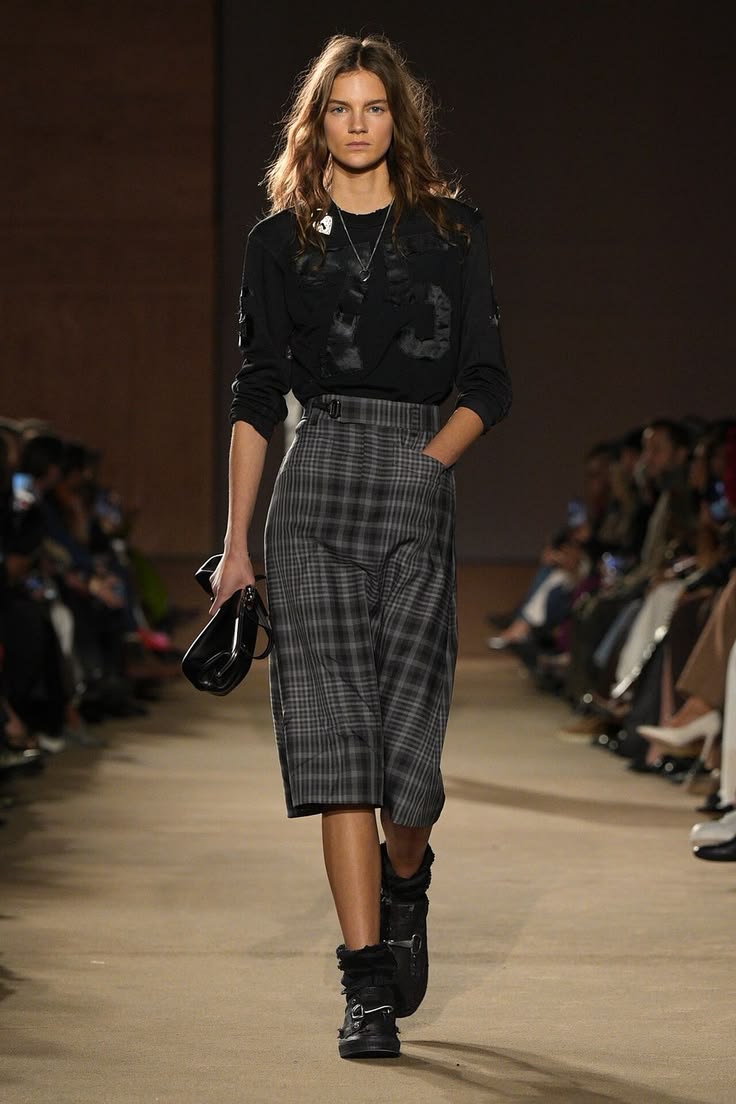 Modelo luciendo una chamarra de cuero negro, un suéter de punto gris de cuello tortuga, bermudas de sastre grises con pinzas y calentadores de lana sobre zapatos oscuros.