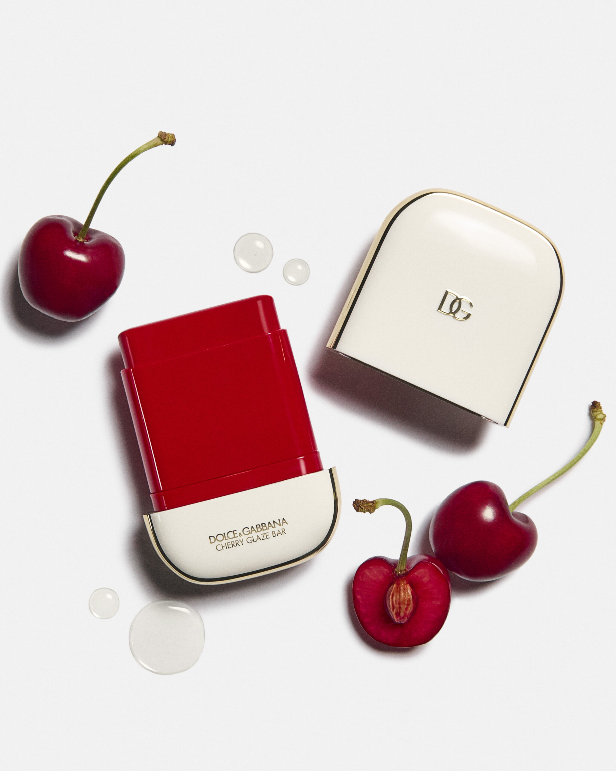 Barra de labios Cherry Glaze abierta junto a su estuche blanco con detalles dorados, rodeada de cerezas frescas enteras y cortadas, y gotas de agua.