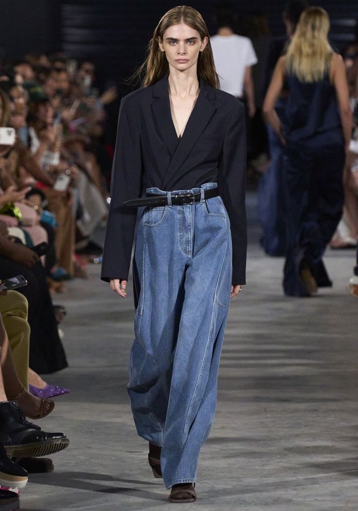 Modelo caminando con un blazer oscuro cruzado por dentro de unos jeans amplios de lavado clásico ajustados con un cinturón negro delgado.