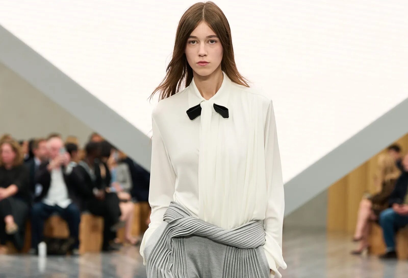 Modelo luciendo una blusa blanca de cuello alto con lazo negro y pantalones grises fluidos con un diseño cruzado y plisado en la cintura.
