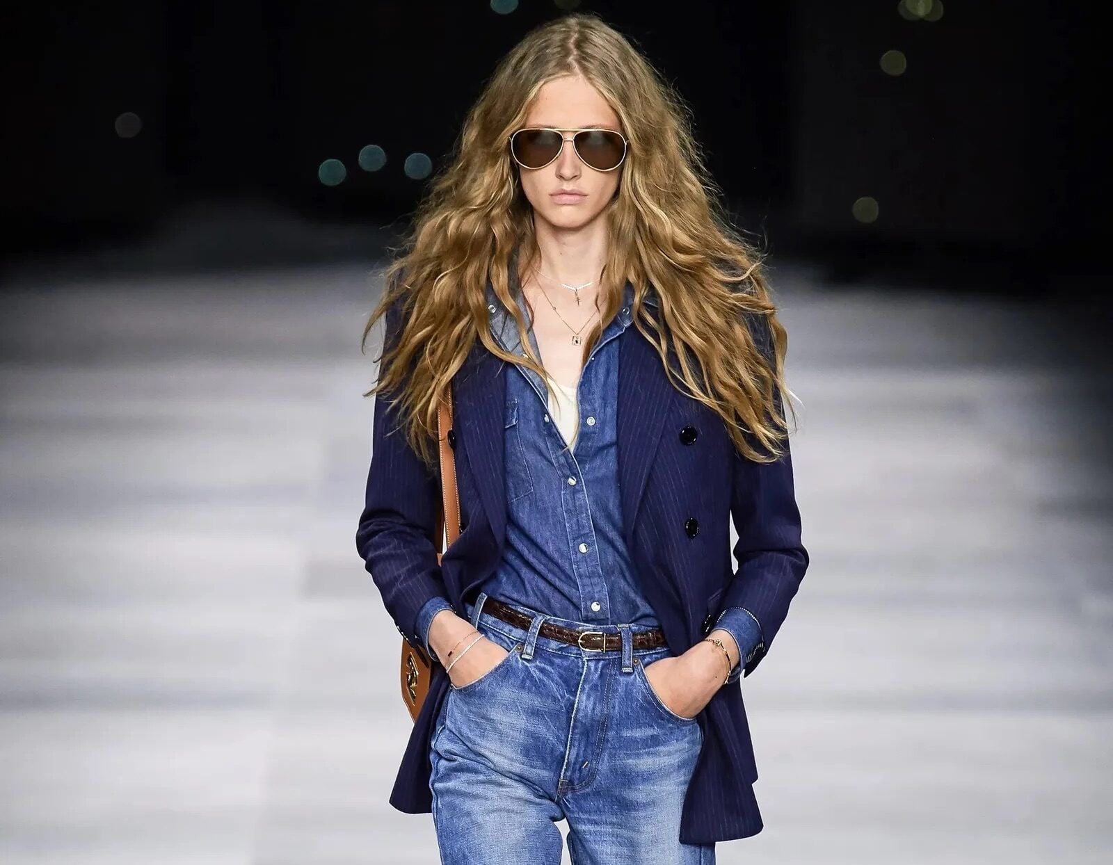 Modelo en pasarela luciendo jeans bootcut de lavado clásico, camisa de mezclilla abotonada, blazer cruzado azul marino, gafas de sol estilo aviador y bolso marrón.