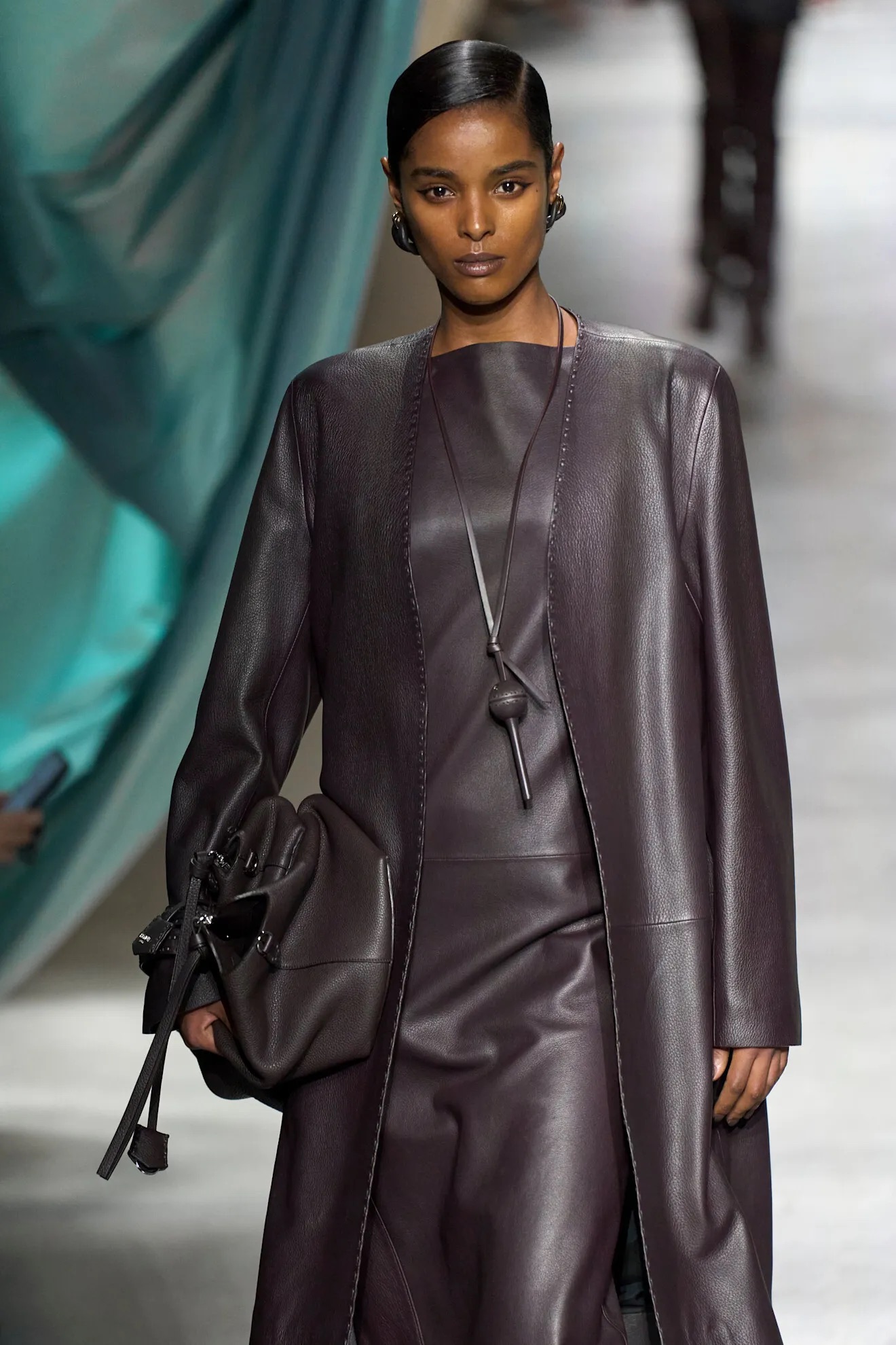 Modelo en pasarela vistiendo un conjunto de cuero marrón oscuro, abrigo largo a juego y un collar colgante largo con un dije cilíndrico de diseño utilitario.