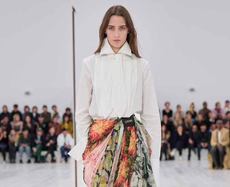 Modelo desfilando con una blusa corta blanca de manga corta con volantes y cuello en V, combinada con pantalones de rayas azules y blancas de talle bajo.