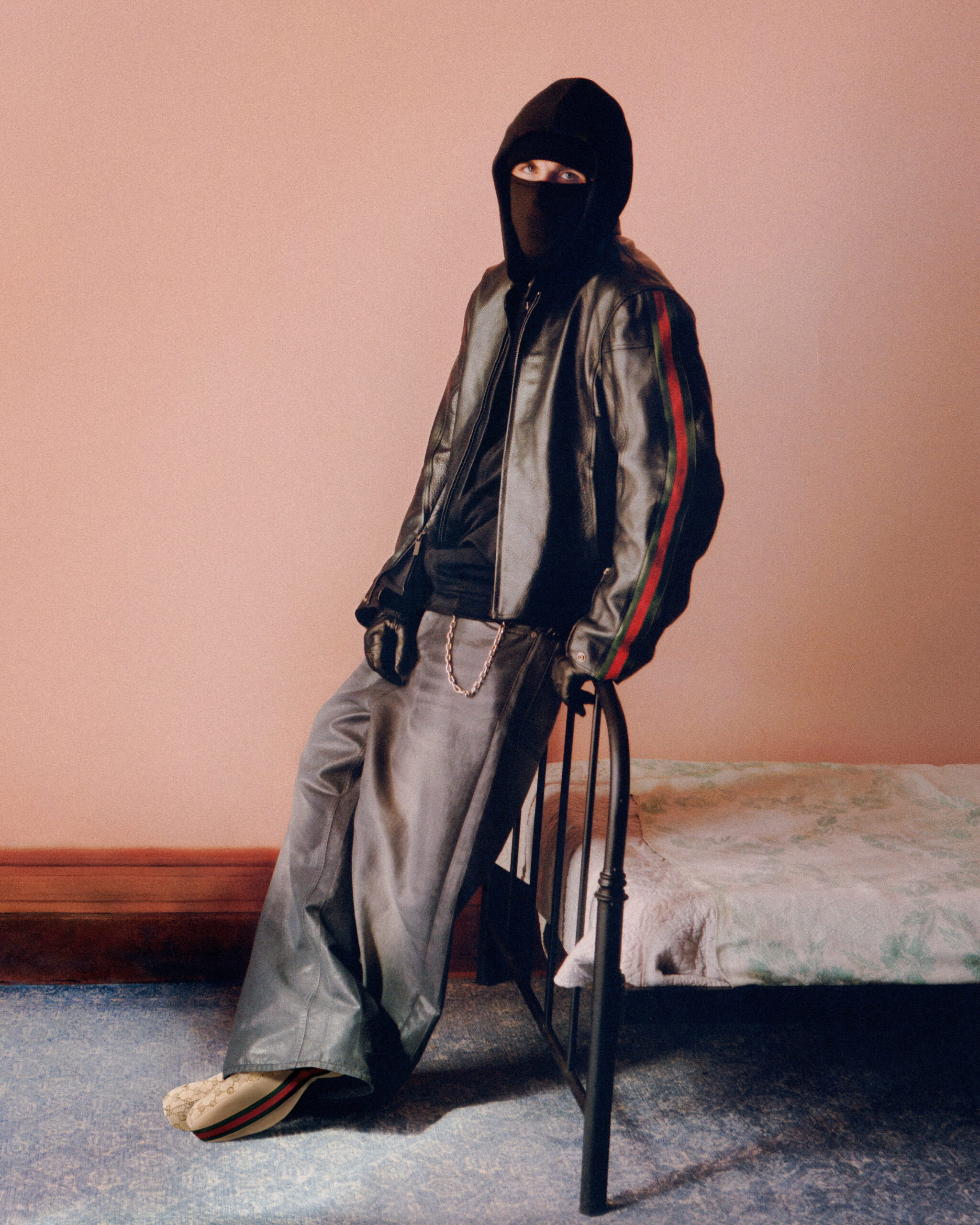 Esdeekid posando junto a una cama de hierro forjado, vistiendo una chamarra de piel negra con bandas laterales rojas y verdes, capucha oscura con pasamontañas y calzado con el monograma de la marca.