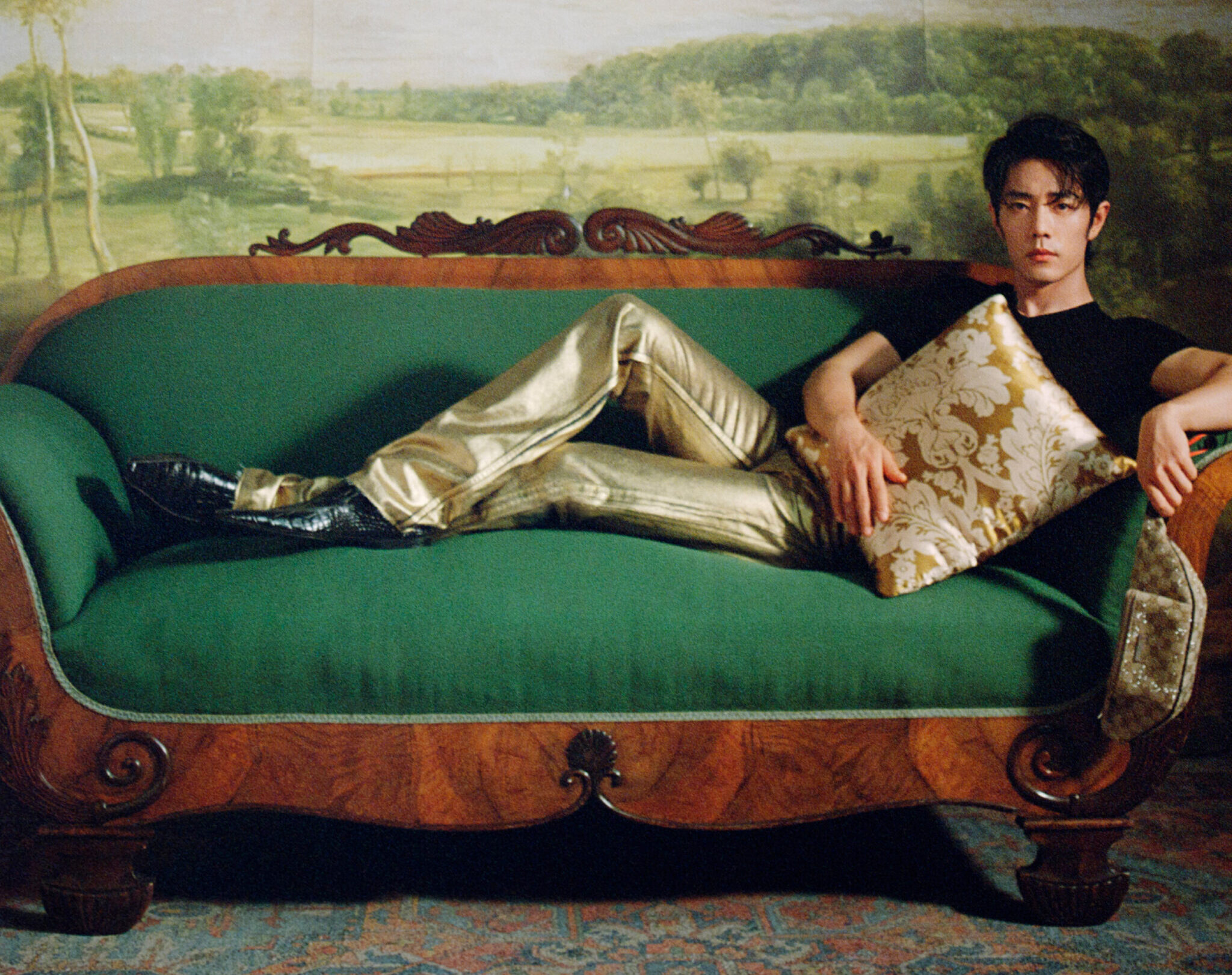 Xiao Zhan recostado relajadamente sobre un sofá verde esmeralda con marco de madera tallada, vistiendo pantalones dorados metalizados y una camiseta negra básica.