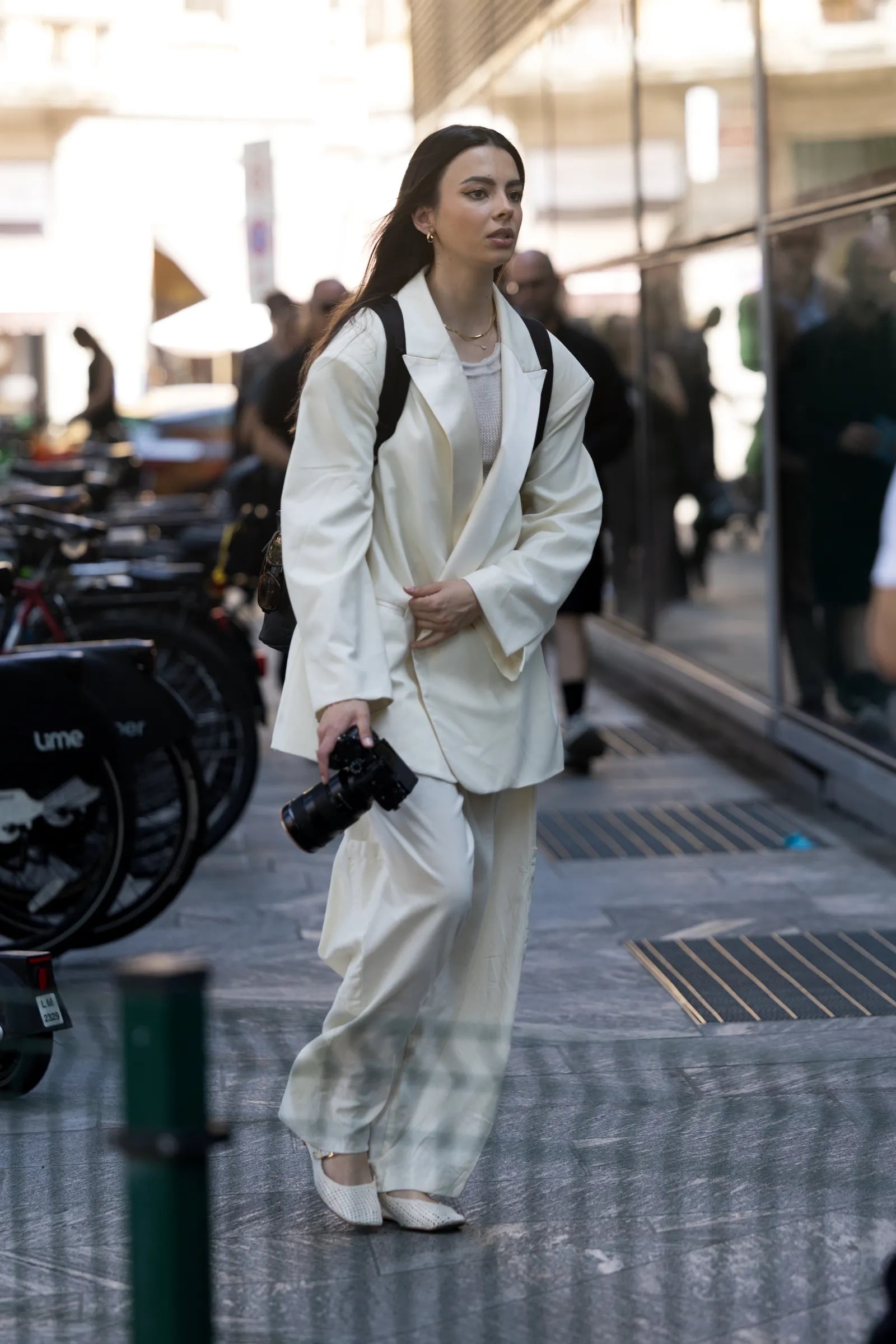 Mujer posando con un traje sastre blanco de corte relajado, mochila, cámara fotográfica y bailarinas slingback texturizadas en color blanco.