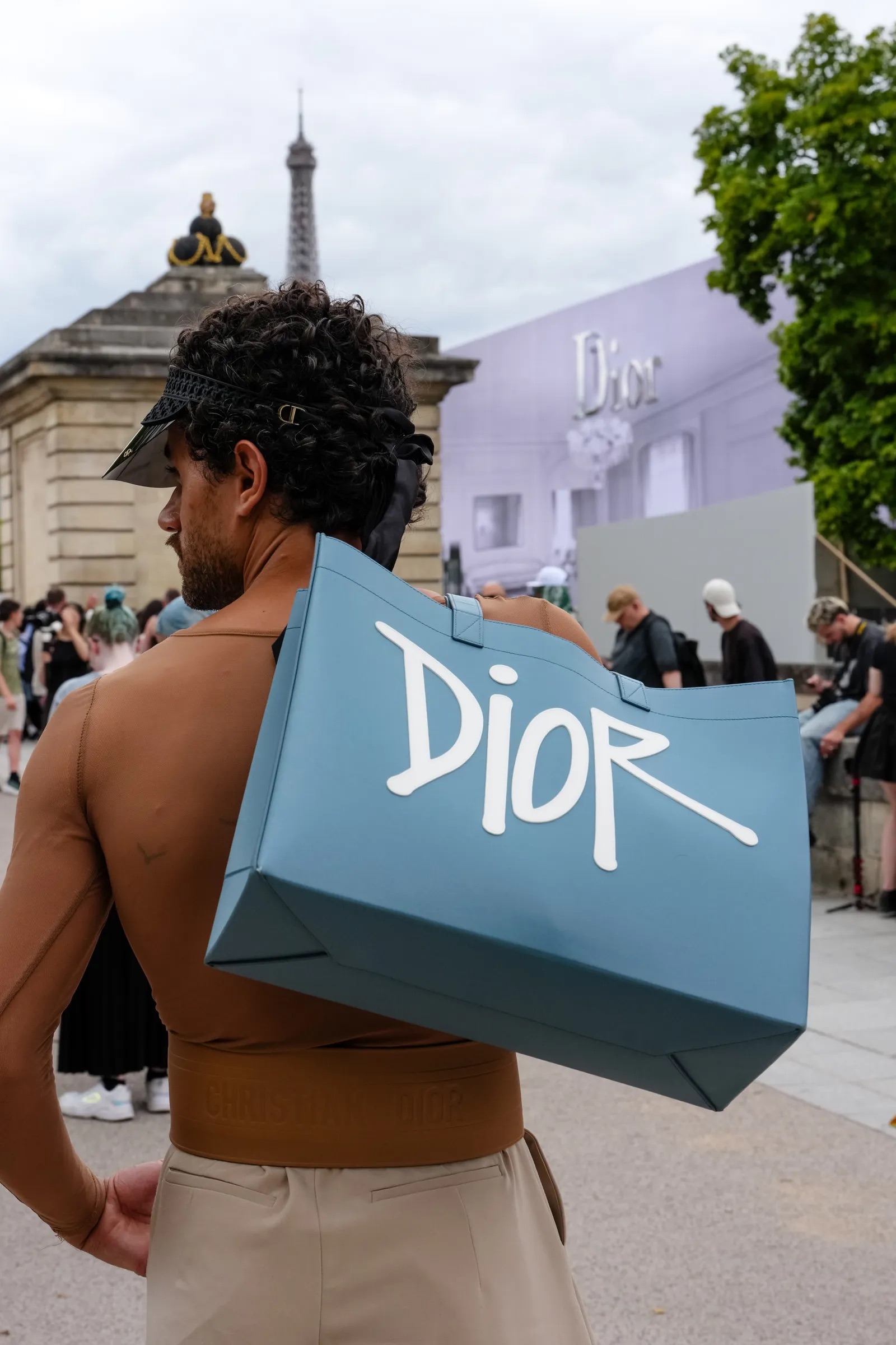 Persona de espaldas con un gran bolso tote azul de Dior frente a un edificio con la Torre Eiffel de fondo