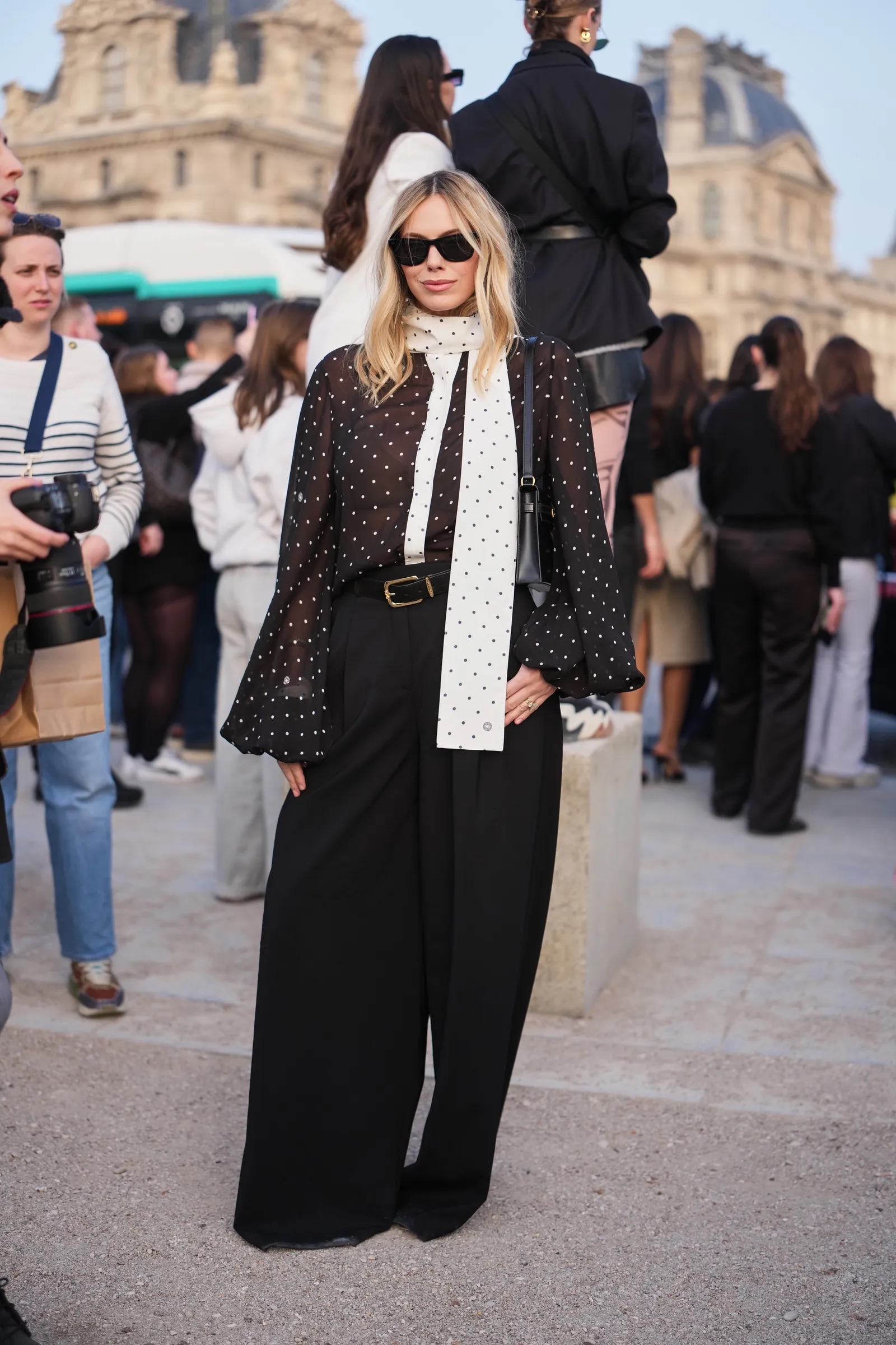 Mujer rubia con gafas de sol, blusa negra translúcida de lunares blancos con pañuelo integrado, pantalones anchos negros y cinturón clásico.