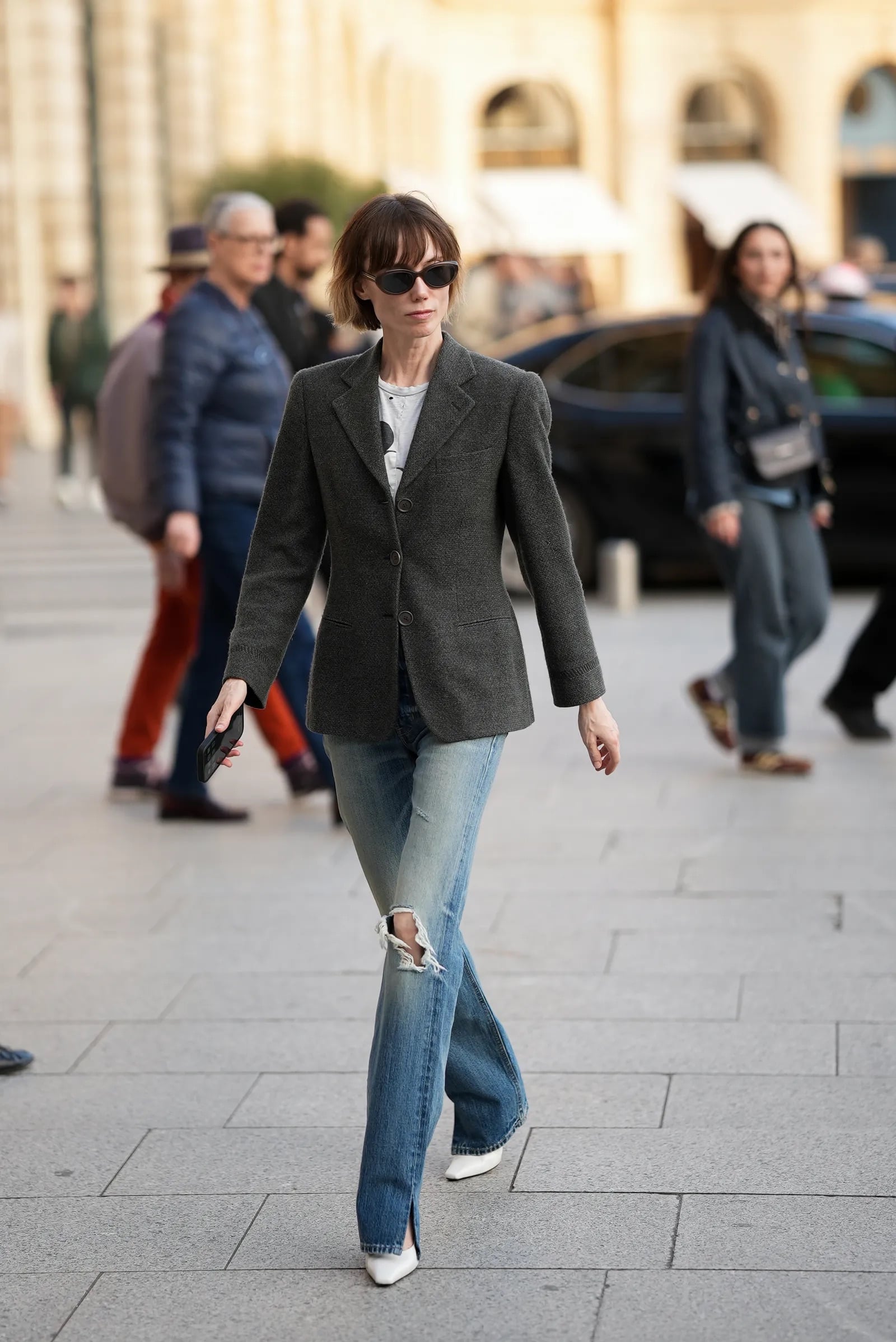 Mujer de perfil con cabello corto caminando por la calle, vistiendo una camiseta estampada, un blazer gris estructurado, jeans ajustados y zapatos de tacón bajo blancos.