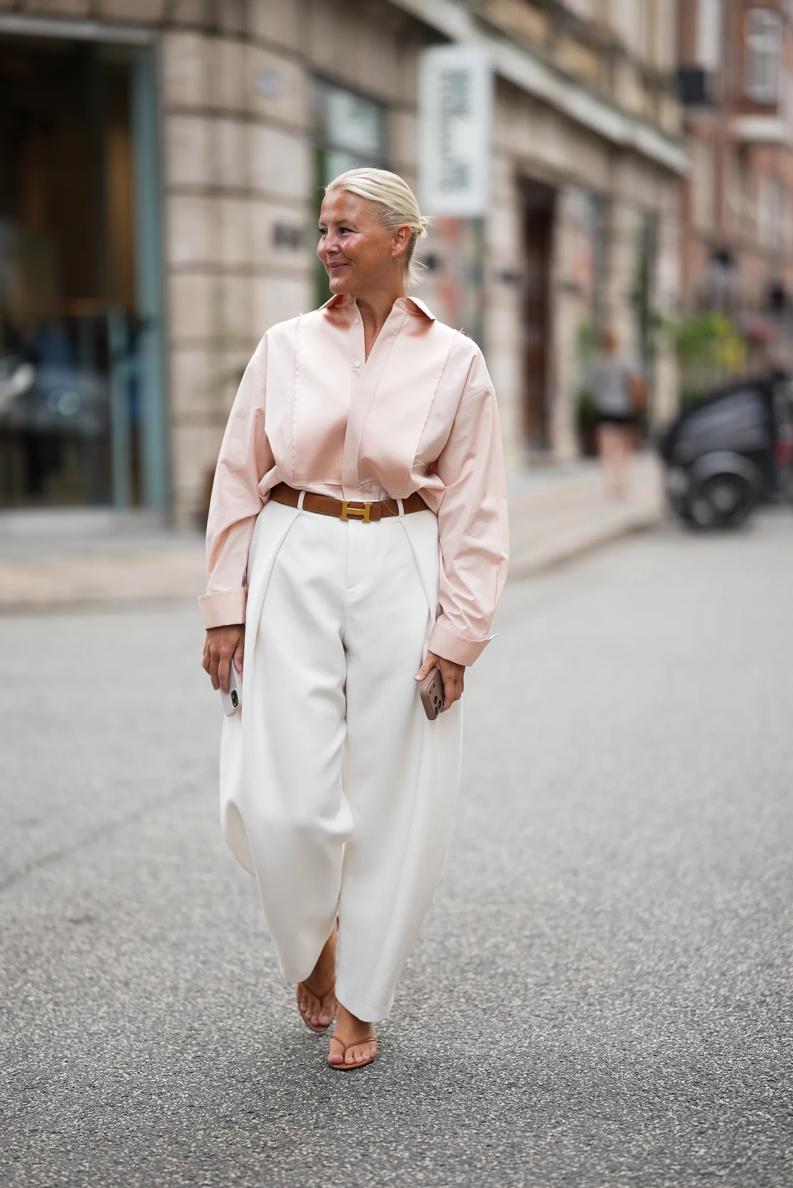 Mujer rubia con camisa rosa claro, pantalones amplios blancos y cinturón marrón en la calle.