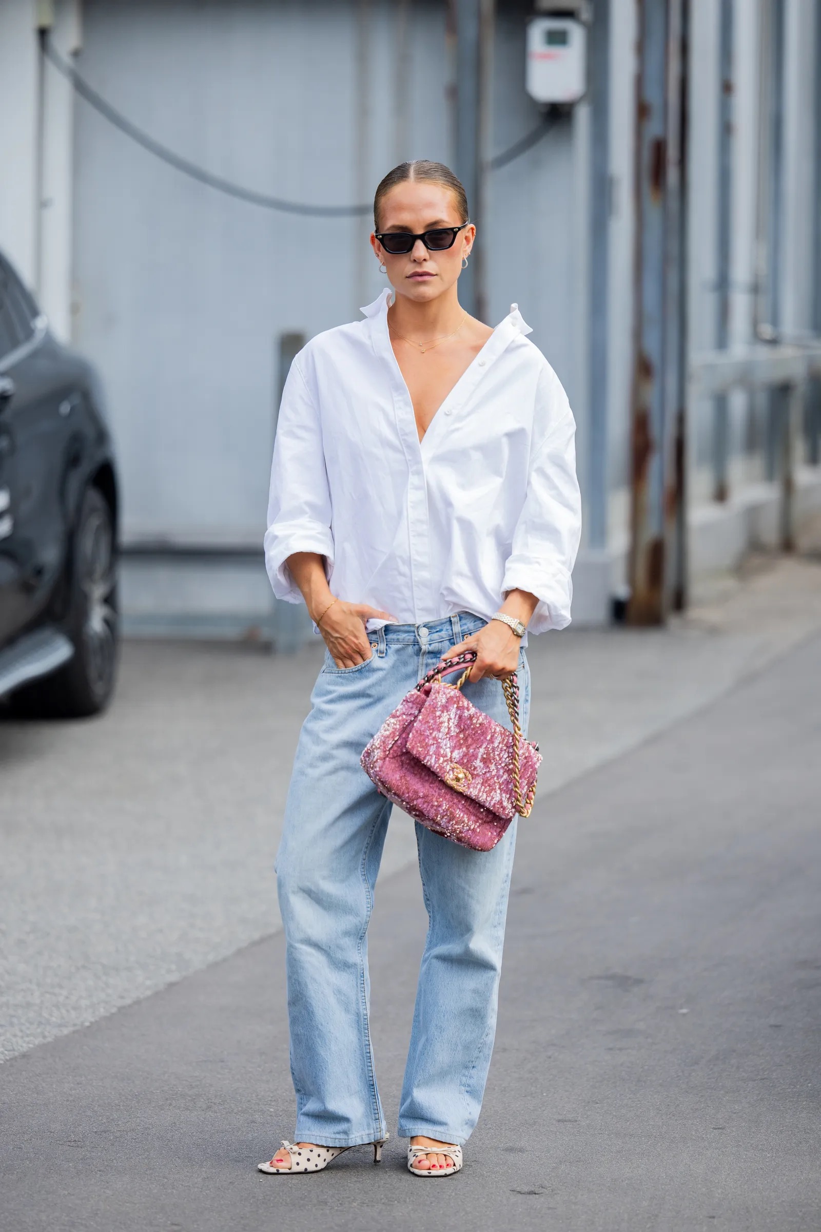 Mujer con camisa blanca oversize, jeans claros y un bolso rosa de tweed brillante en la calle.