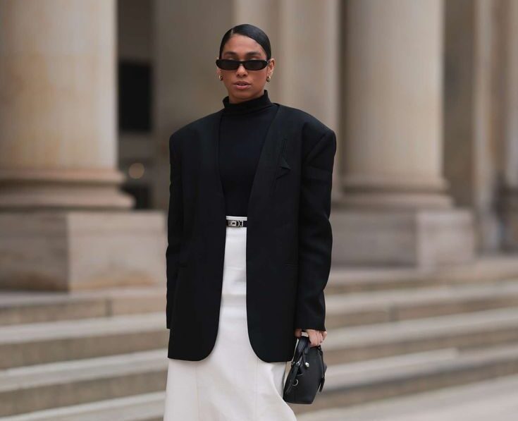 Persona con cuello alto negro, blazer oversize estructurado, falda midi blanca con cinturón fino y zapatos de tacón destalonados.