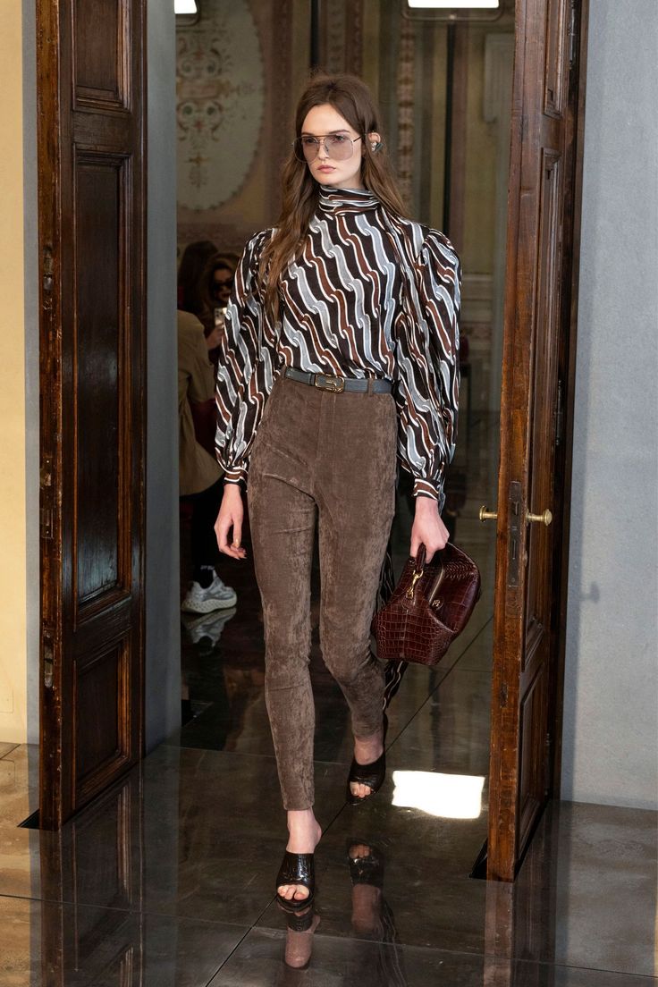 Modelo con blusa de cuello alto estampada y leggings ajustados de pana marrón, combinados con un bolso de mano de piel texturizada.