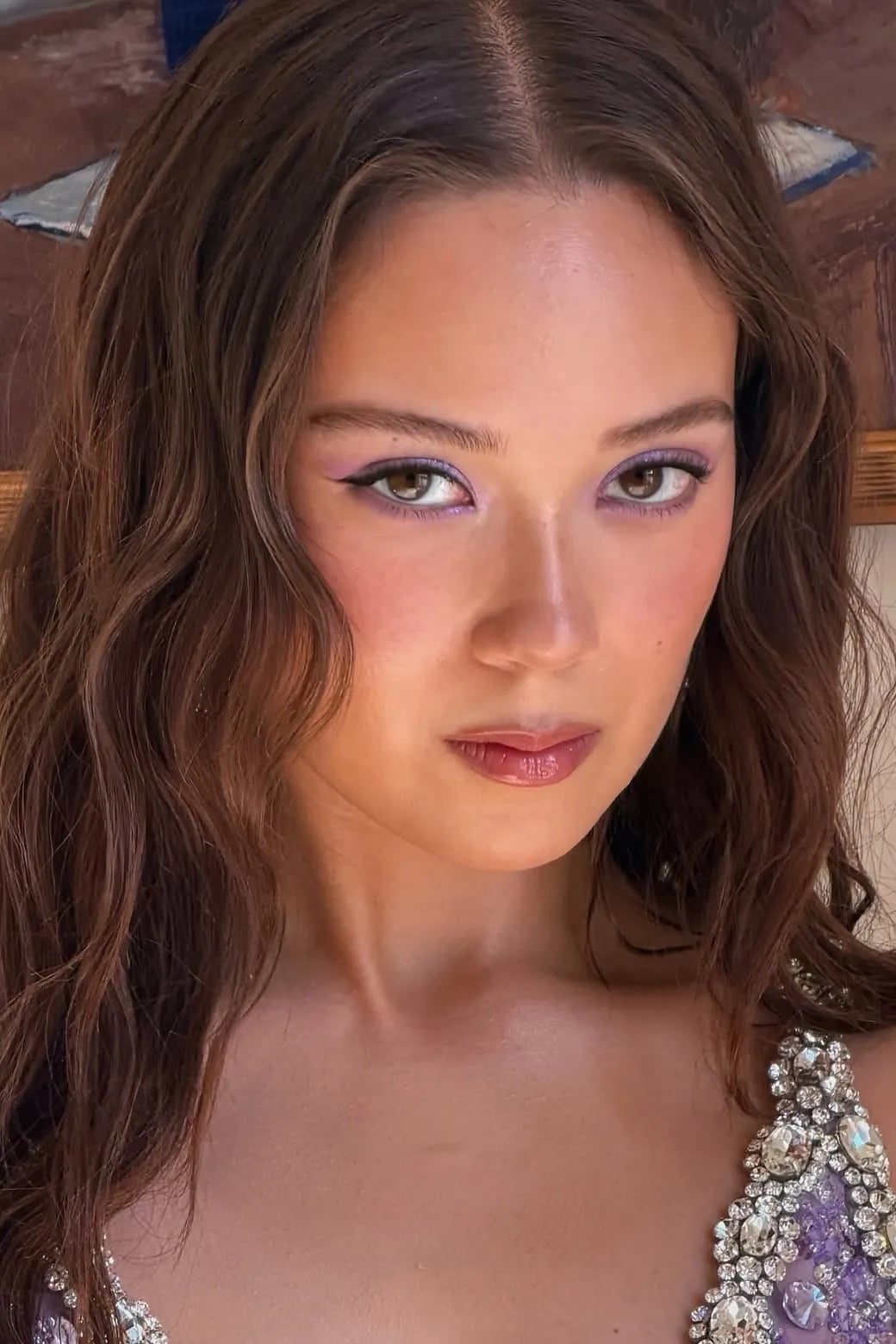 Primer plano de Laufey con cabello ondulado, luciendo sombras y delineado en tonos morados suaves con un vestido brillante de pedrería.