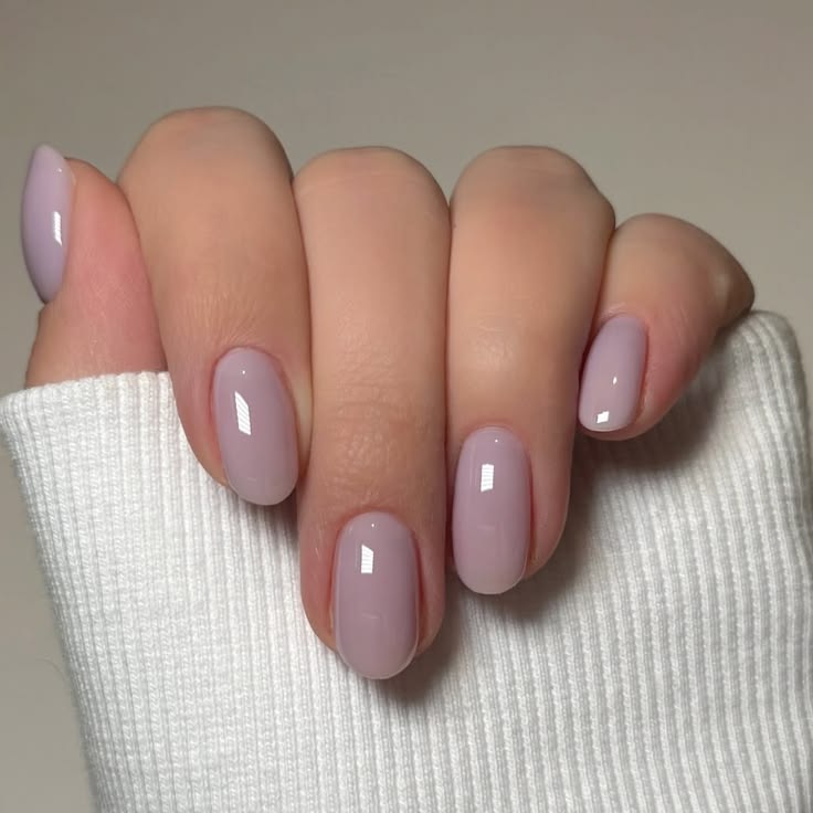 Mano con uñas cortas de forma cuadrada y suave, con una manicura en tono lavanda cremoso y brillante, junto a una prenda clara.