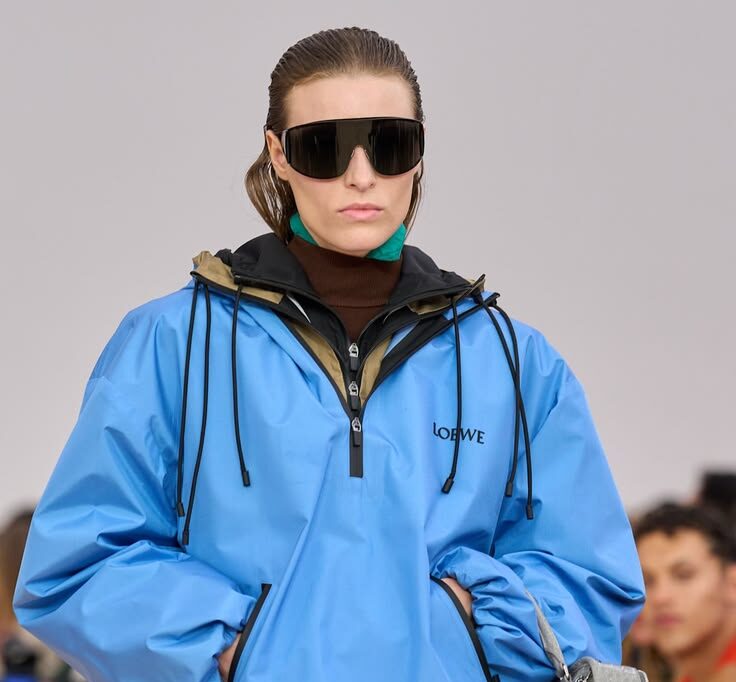 Modelo con gafas de sol oscuras luciendo una chamarra deportiva tipo cortavientos en color azul brillante con capucha, sobre capas de suéteres y detalles en tonos tierra y verde.