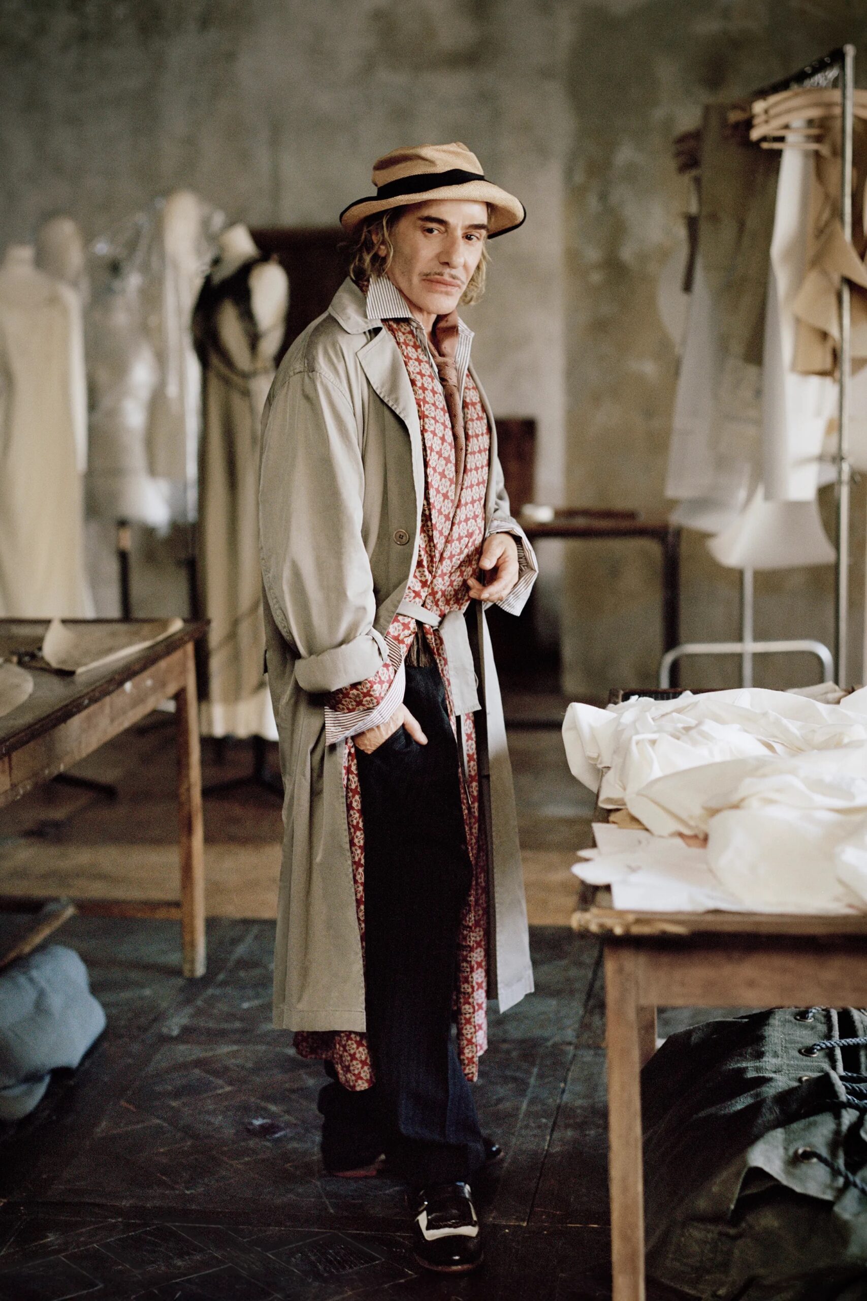 John Galliano en su taller de alta costura, vistiendo gabardina clara, sombrero y un pañuelo estampado entre maniquíes y telas.