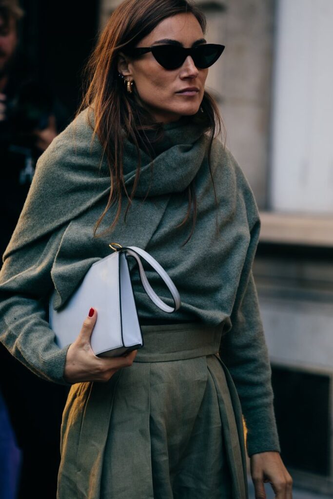 Modelo luciendo una prenda de punto en tono verde con el cuello drapeado a modo de bufanda o pañuelo, gafas de sol oscuras y un bolso de mano blanco.