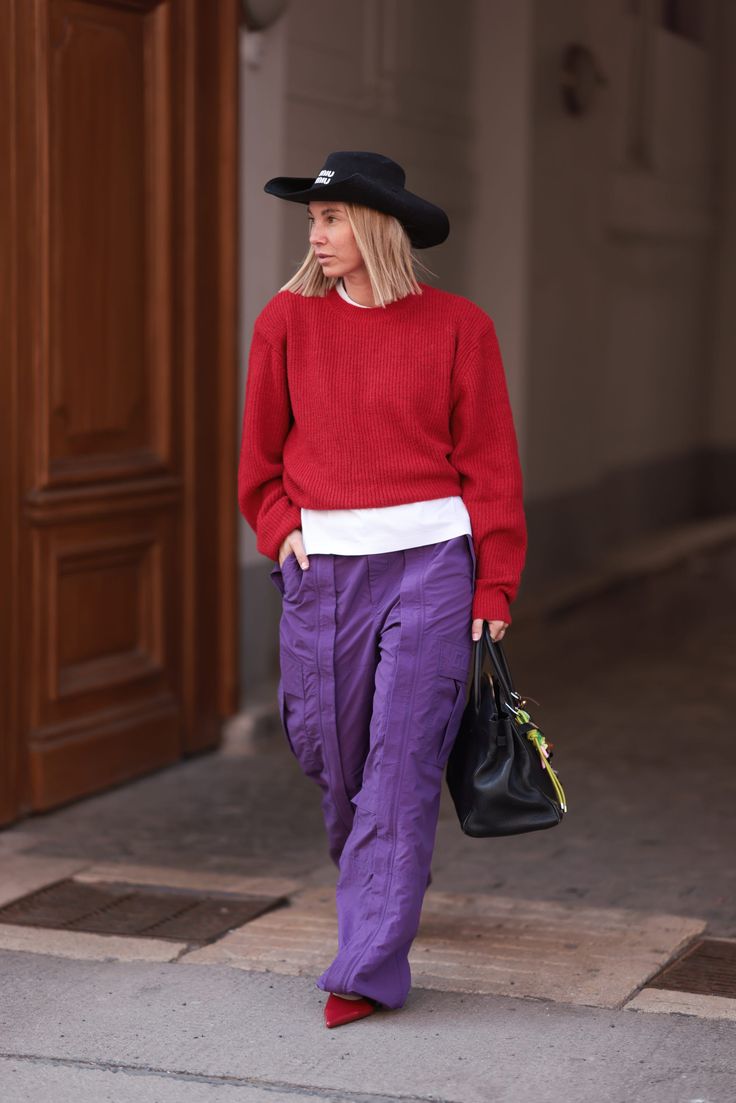 Mujer caminando por la calle luciendo un suéter de punto rojo, pantalones cargo morados amplios, sombrero negro y zapatos rojos.
