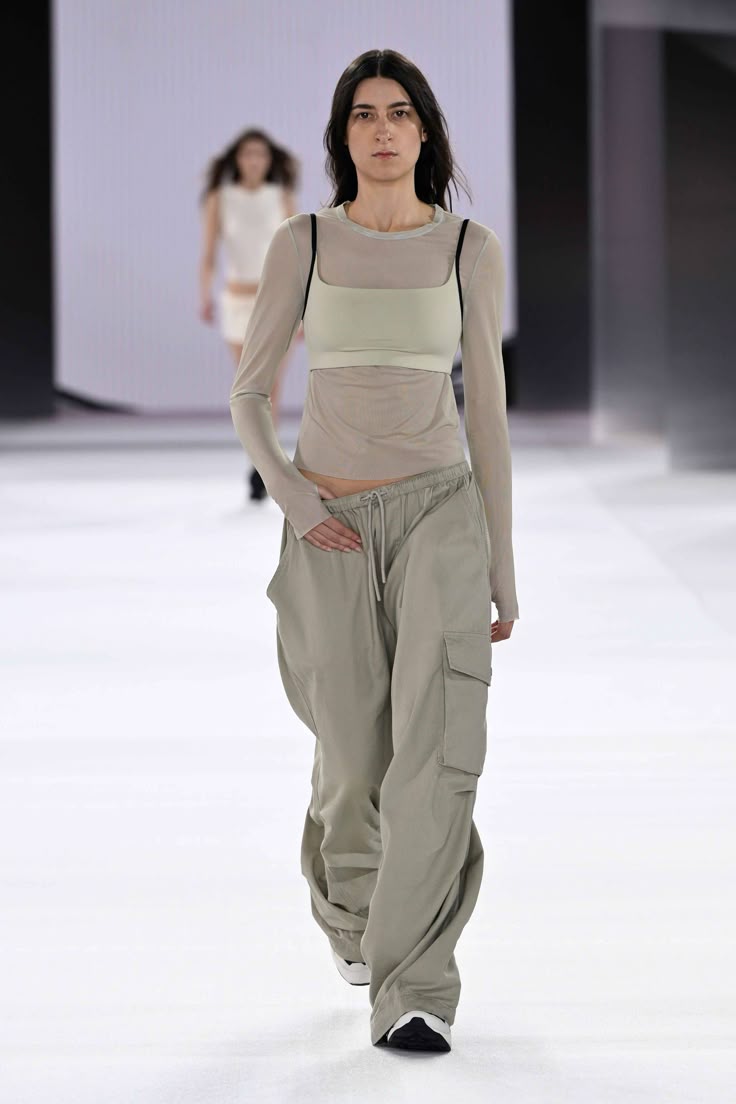 Modelo caminando en la pasarela con un top translúcido de manga larga en tono neutro, un sostén a juego por debajo y pantalones cargo amplios de talle bajo en color gris claro.