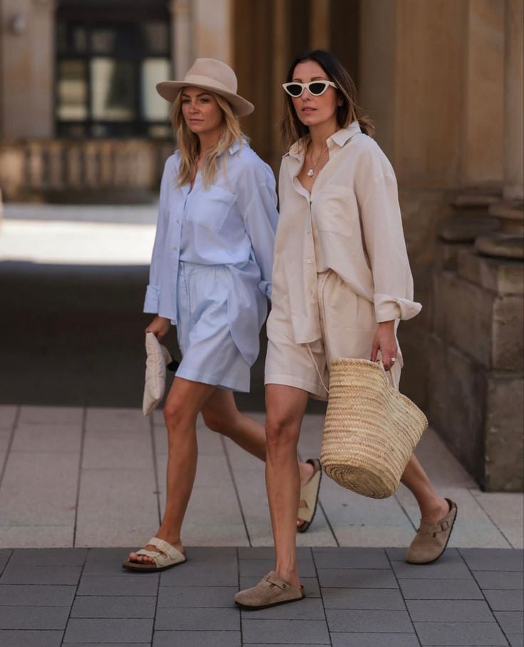 Dos mujeres caminando por la calle con conjuntos monocromáticos de camisa y bermudas, acompañados de sandalias planas y zuecos, una con sombrero y la otra con bolsa de mimbre.