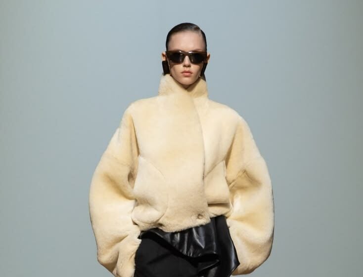 Modelo con gafas de sol luciendo una voluminosa chaqueta corta de pelo sintético en tono marfil sobre pantalones negros de sastre.