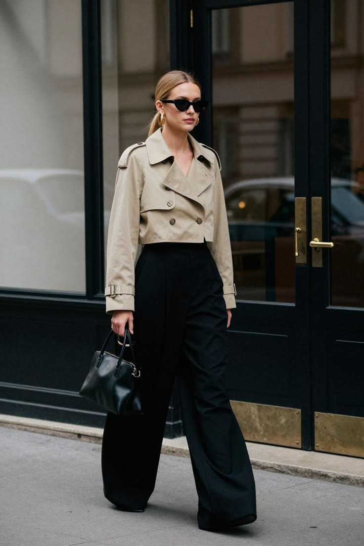Modelo caminando en la calle con una gabardina corta en tono beige, pantalones negros de pernera ancha, gafas de sol oscuras y un bolso de mano negro.