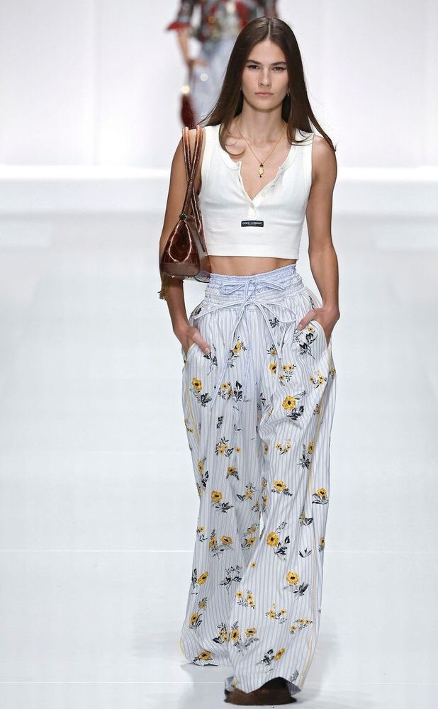 Modelo en pasarela con un top corto blanco de tirantes con botones frontales, combinado con pantalones fluidos de rayas y estampado floral en tonos amarillos.