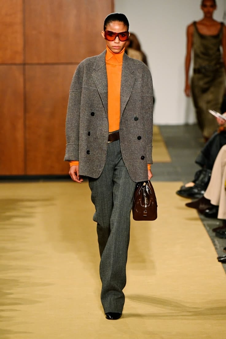 Modelo luciendo un suéter de cuello alto en tono naranja brillante bajo un blazer gris de tweed, pantalón de sastre a juego y gafas de sol con montura roja.