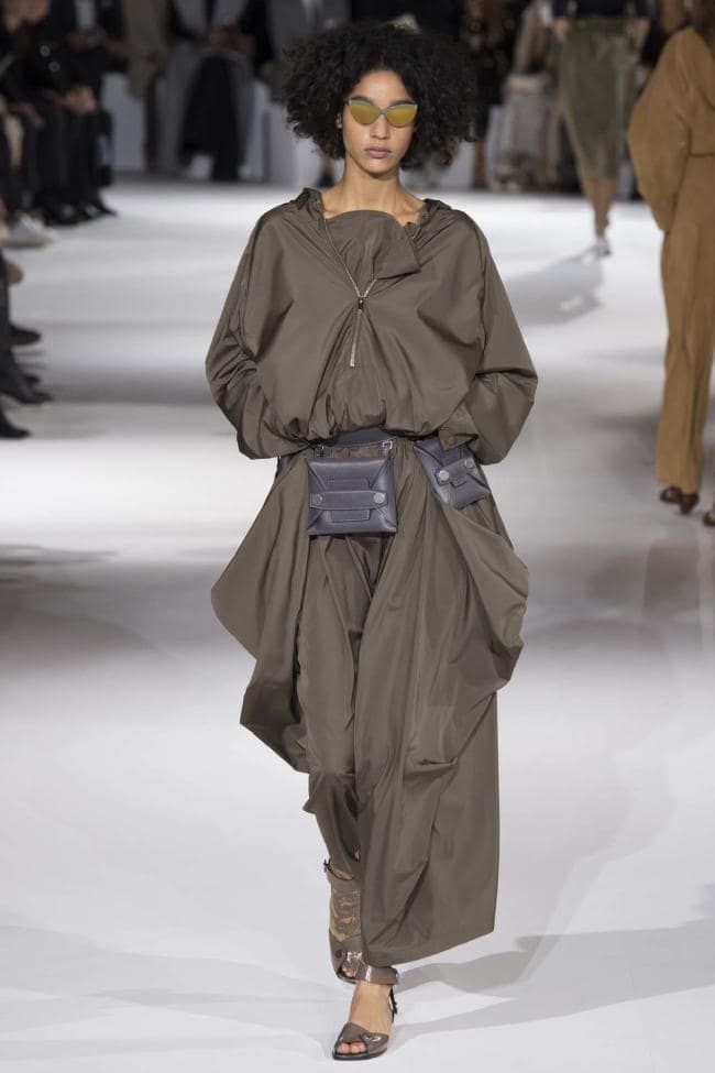Modelo de rizos oscuros desfilando con un conjunto impermeable en tono verde militar compuesto por una chamarra con cierre y falda larga fluida, complementado con un cinturón tipo cangurera.