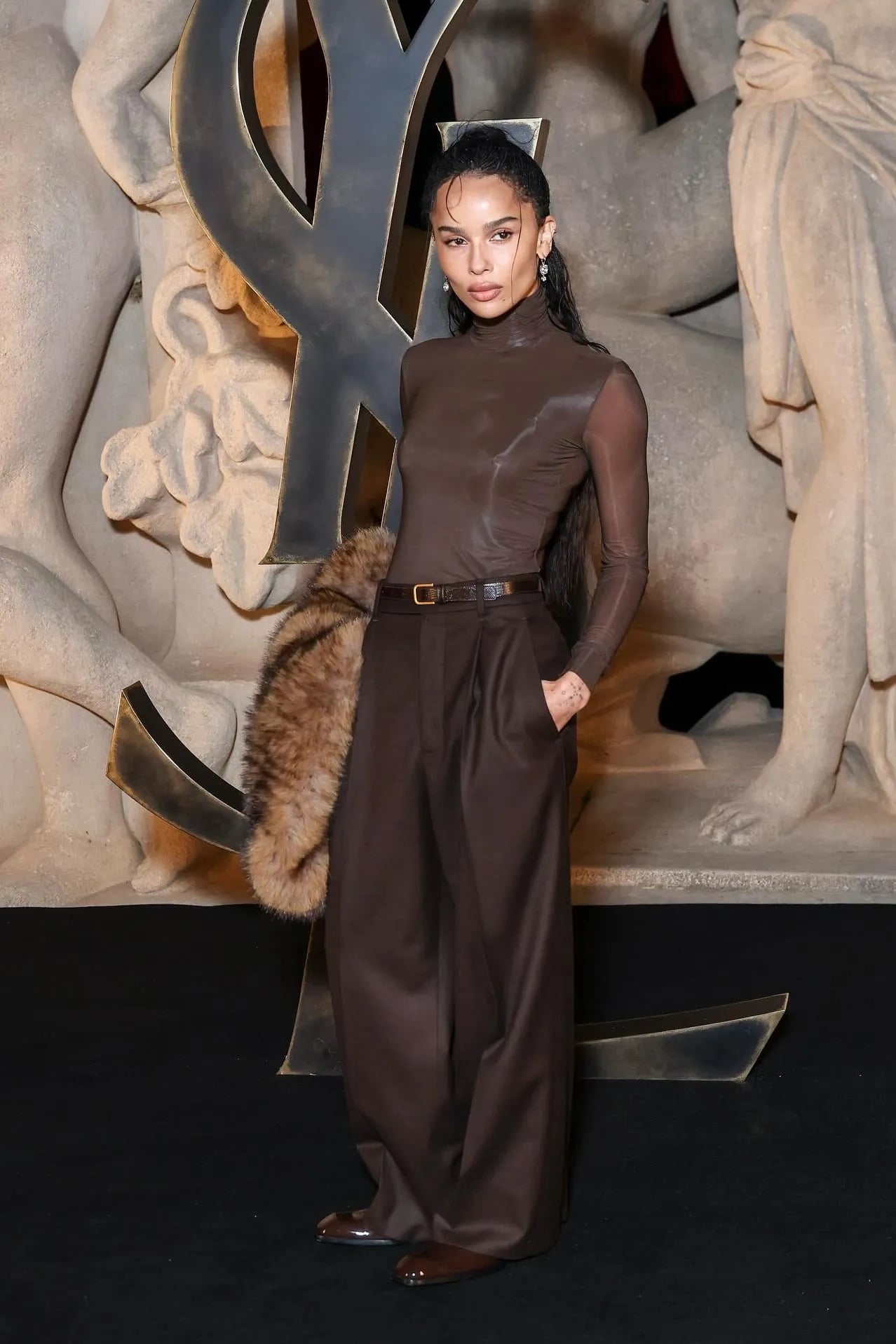 Zoë Kravitz posando con un body ajustado marrón de cuello alto, pantalones de sastre anchos y oscuros, y un accesorio de piel.