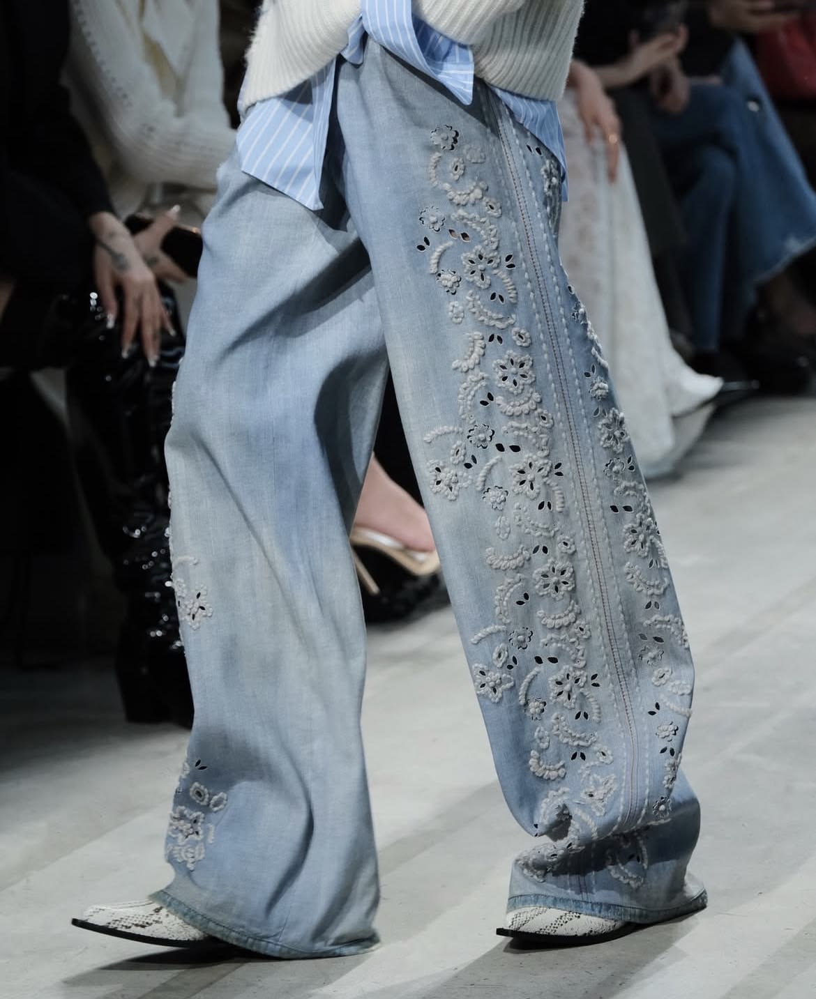Pantalón de mezclilla claro decorado con bordados florales y perforaciones (broderie anglaise) en la pierna, acompañado de una camisa a rayas y suéter de punto