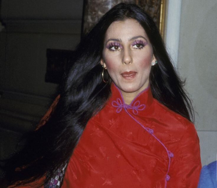 Cher en 1975 con la melena larga suelta, maquillaje dramático en tonos morados en los ojos y una prenda roja de cuello alto con detalles de pasamanería.