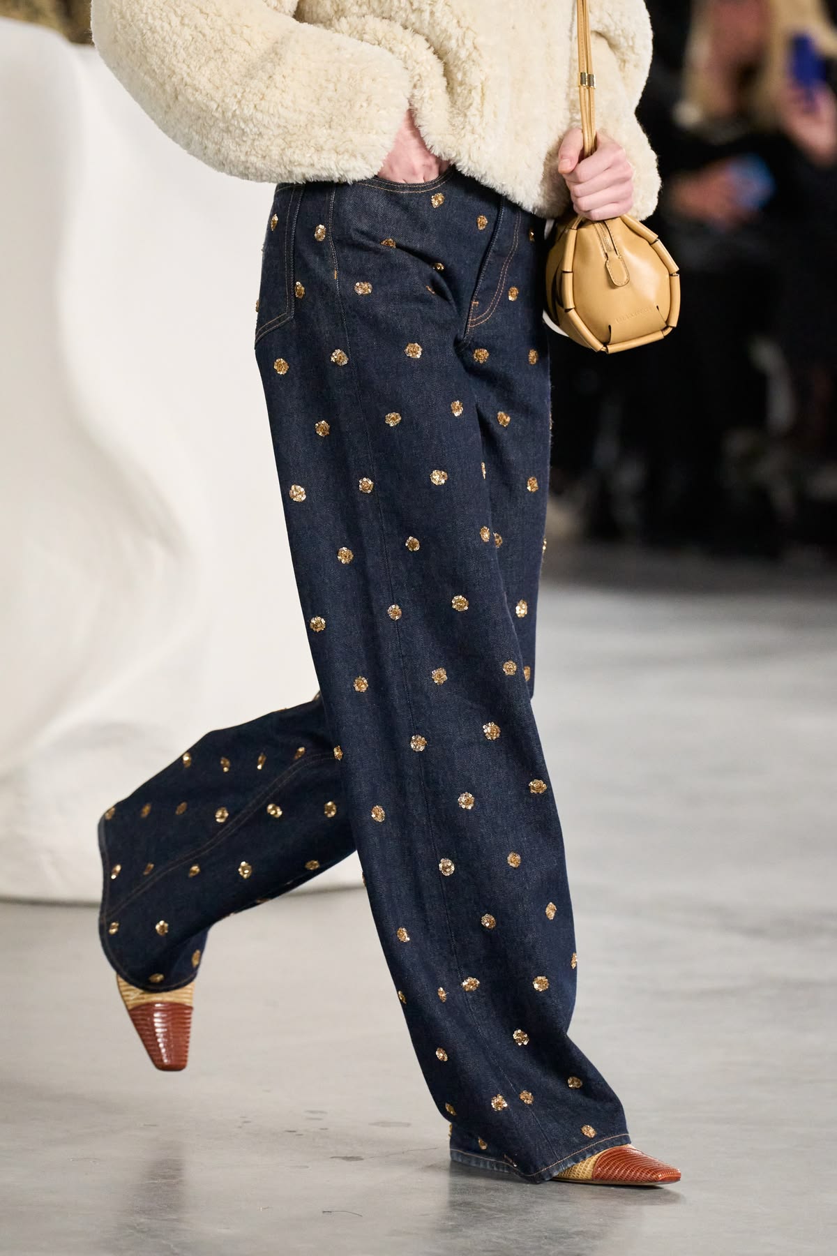 Detalle de jeans oscuros de corte recto repletos de pequeños apliques brillantes y pedrería, combinados con una chaqueta de borrego sintético en tono crema
