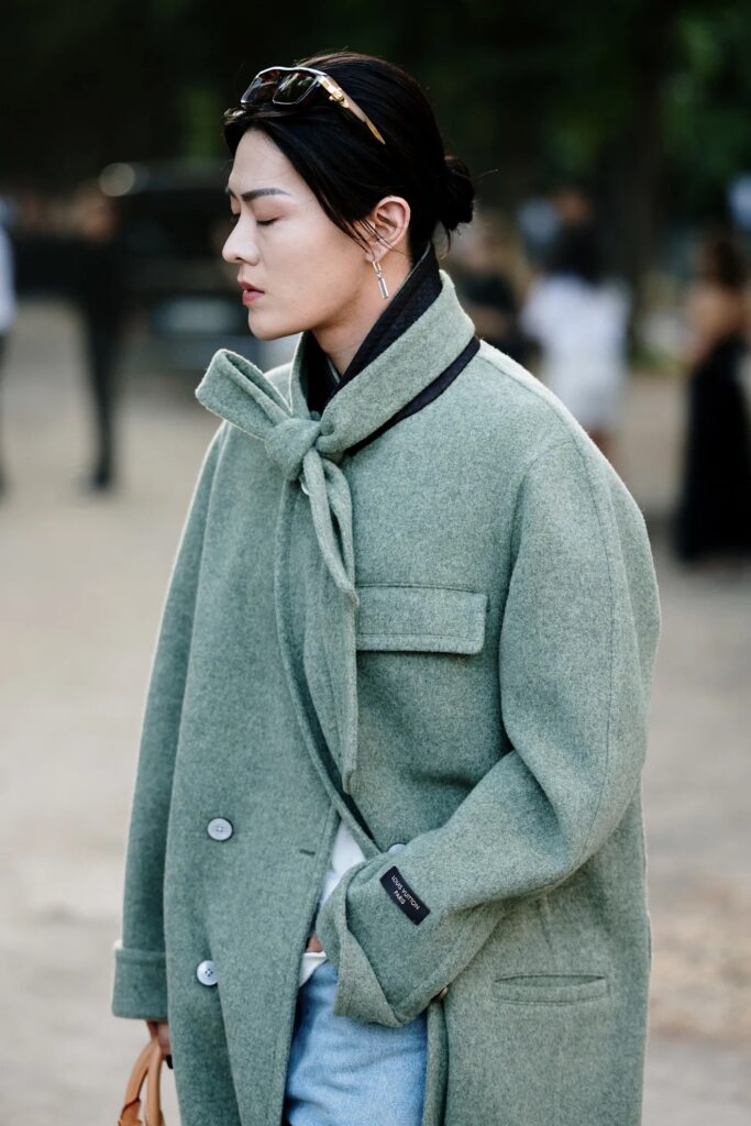 Modelo de perfil con gafas de sol sobre la cabeza luciendo una chaqueta verde oliva estructurada con detalle de cuello tipo pañuelo anudado.