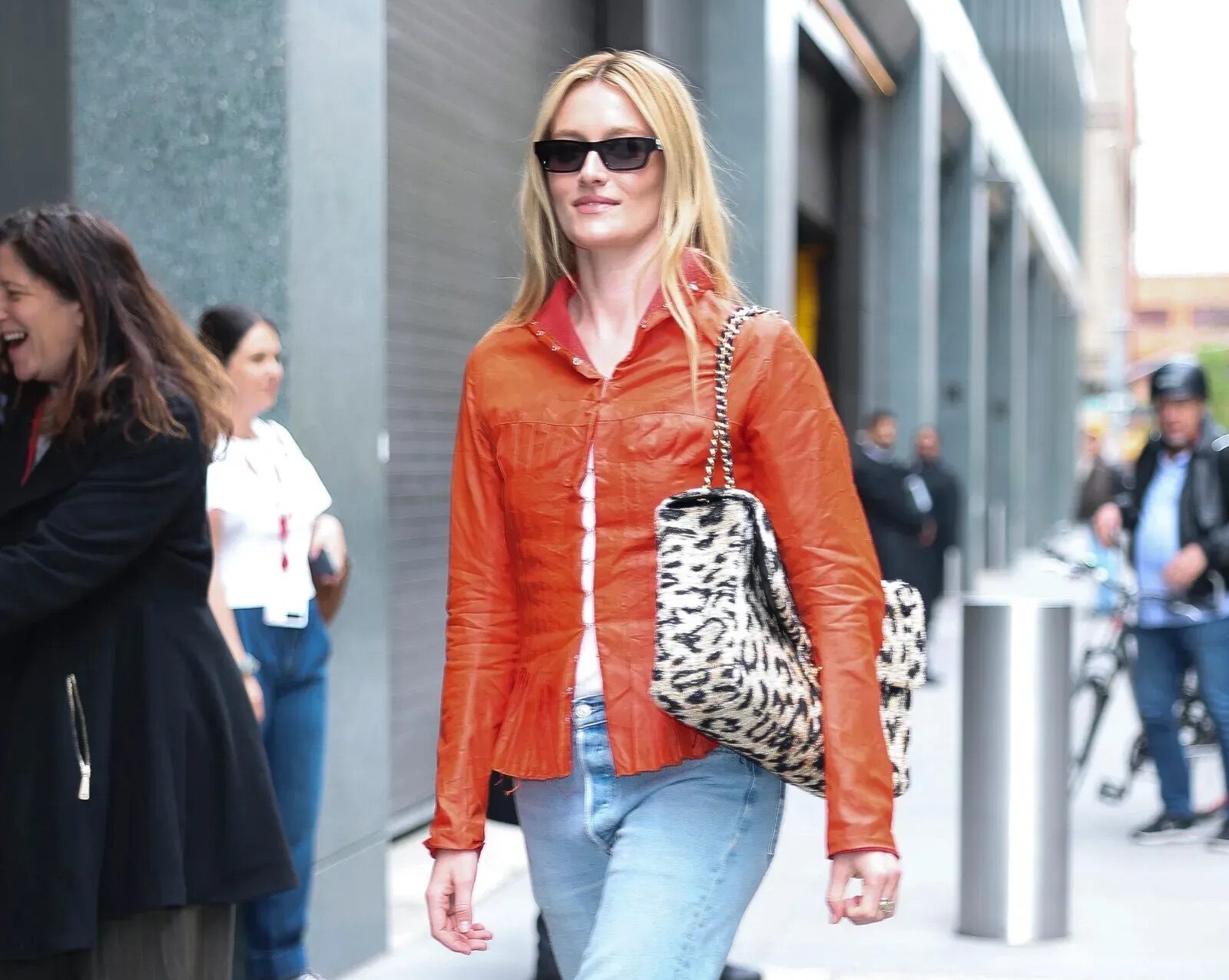 Mujer rubia caminando por la calle con gafas de sol, una chamarra naranja de textura ligera, jeans pitillo rectos y zapatos de tacón bicolor con bolso estampado de leopardo.