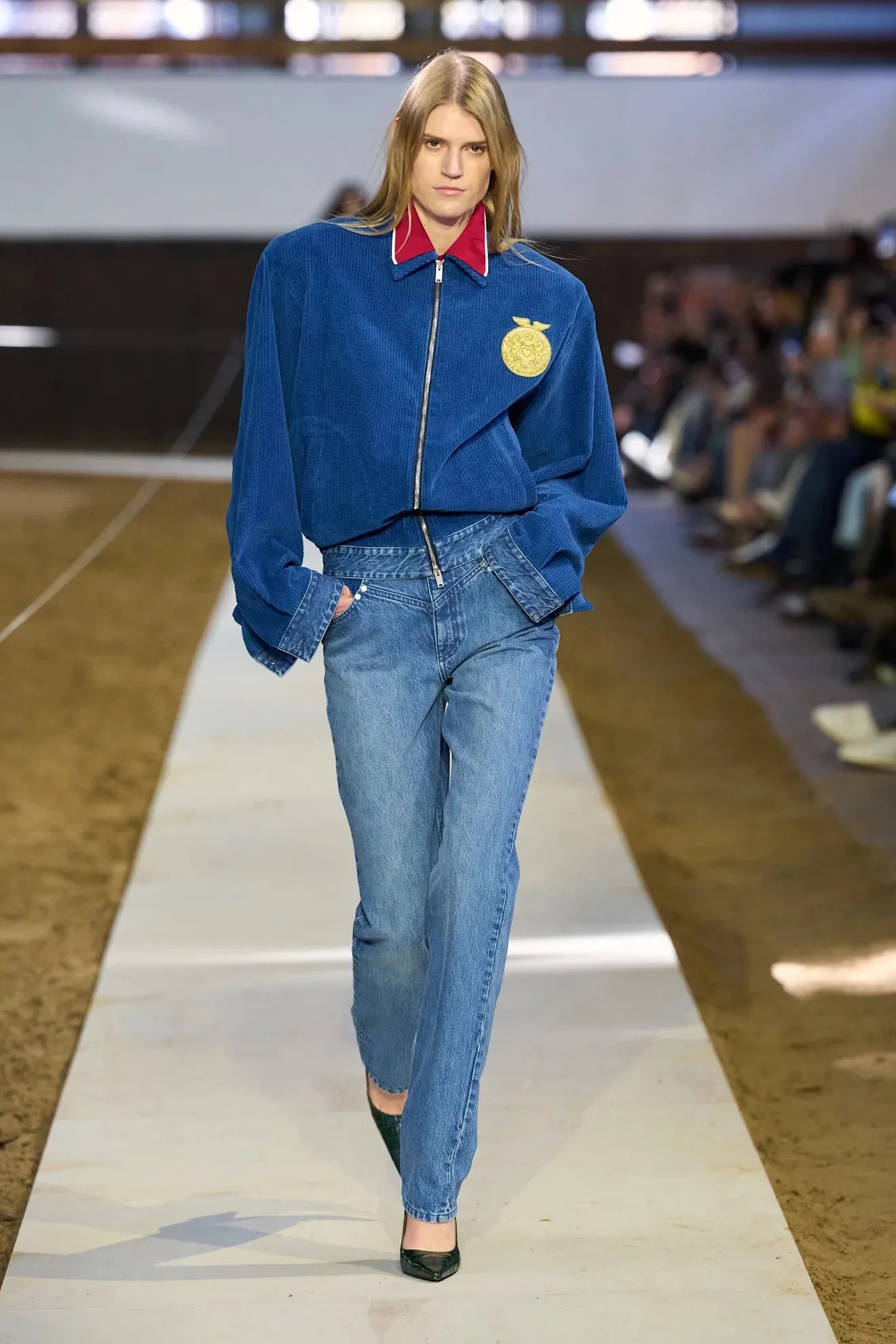 Modelo desfilando en pasarela con una chamarra azul de pana con cuello camisero rojo, jeans pitillo de lavado clásico y zapatos de tacón oscuro.