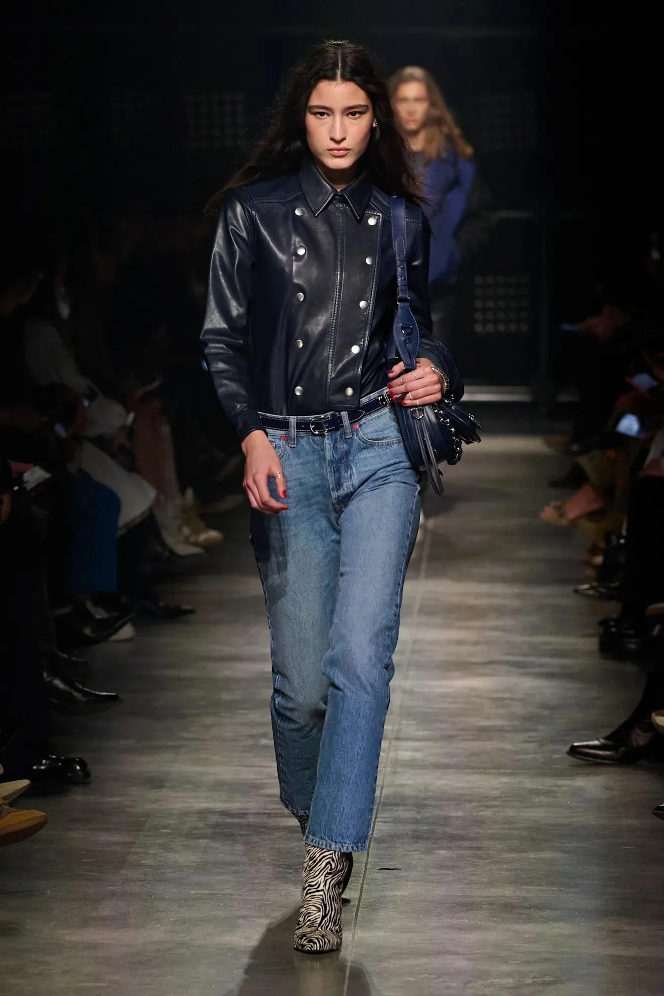 Modelo caminando con una camisa de piel sintética azul marino abotonada, jeans pitillo clásicos, un bolso de hombro a juego y botines estampados con motivo animal.