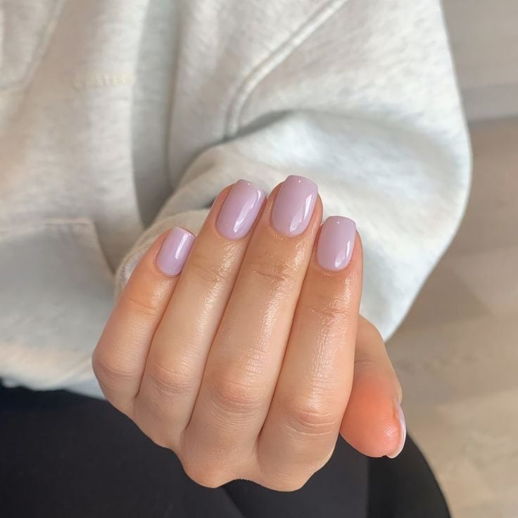 Mano con manicura lavanda clara y translúcida en uñas cortas y cuadradas, luciendo una sudadera gris jaspeada.