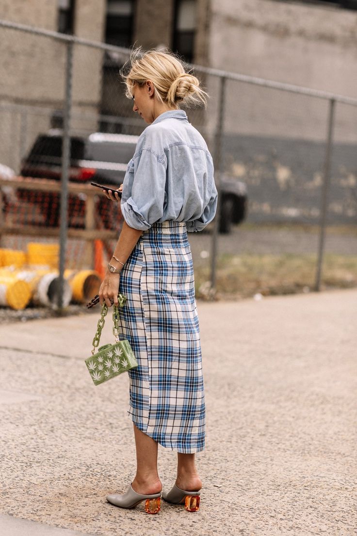 Mujer de espaldas con el cabello recogido, vistiendo una camisa de mezclilla clara ligeramente abombada, una falda midi con estampado de cuadros en tonos azules y blancos, y zapatos destalonados con tacón geométrico.
