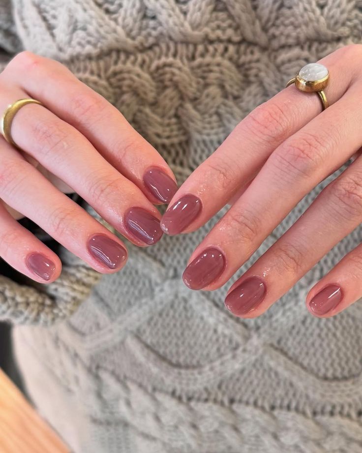Manos entrelazadas luciendo un esmalte de uñas corto en un tono rosa malva profundo y cremoso, con anillos de piedra natural de tonos tierra.