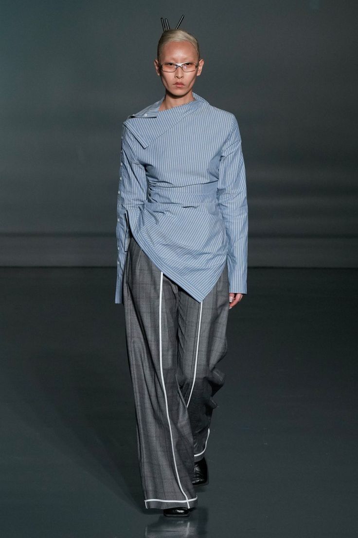 Modelo en pasarela luciendo una camisa asimétrica de rayas azules con cuello cruzado y drapeado, combinada con pantalones de sastre grises de pernera ancha con ribetes blancos.