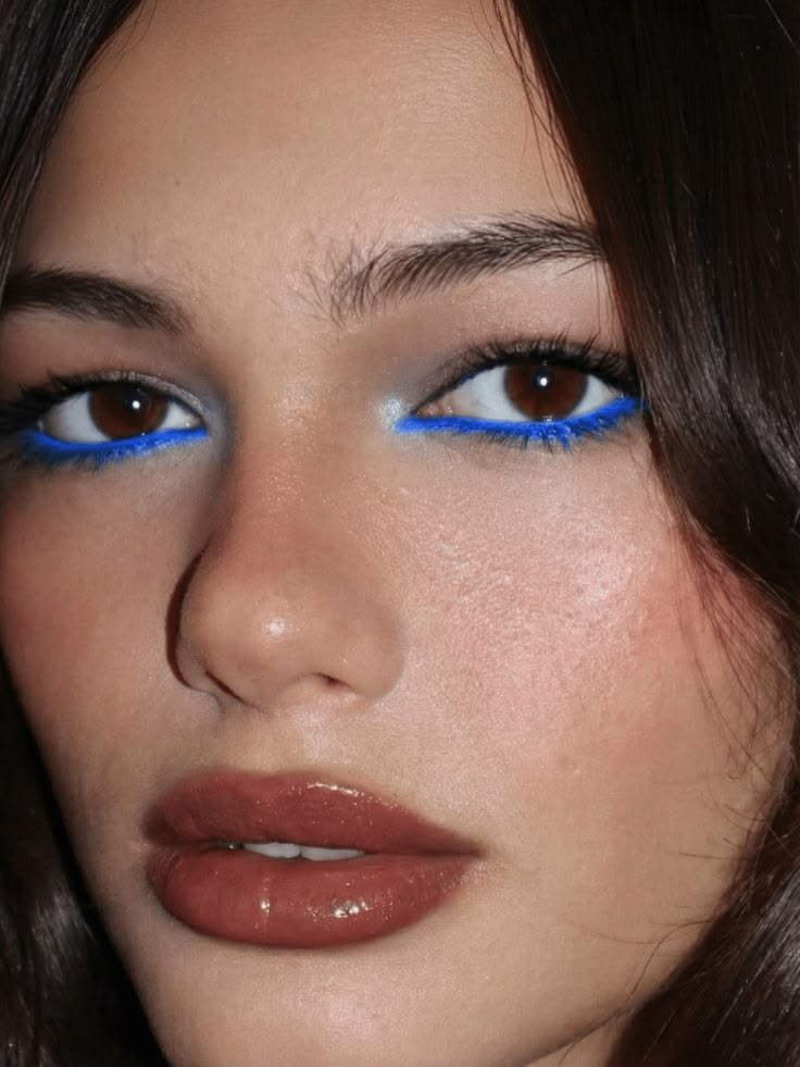 Primer plano de unos ojos castaños delineados en la línea de agua inferior con un tono azul eléctrico intenso y labios en acabado gloss marrón.