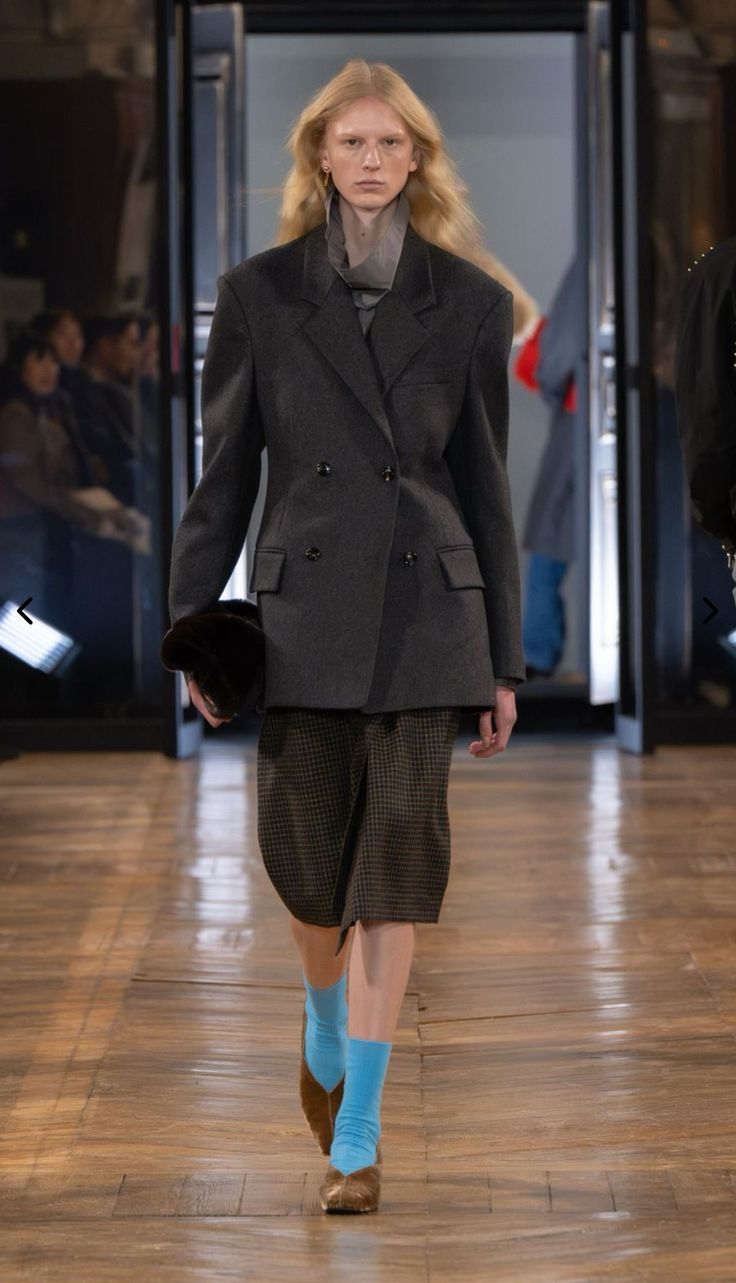 Modelo con cabello rubio desfilando con un abrigo de botonadura doble en tono gris oscuro, falda midi de cuadros, calcetines azules y zapatos planos marrones.