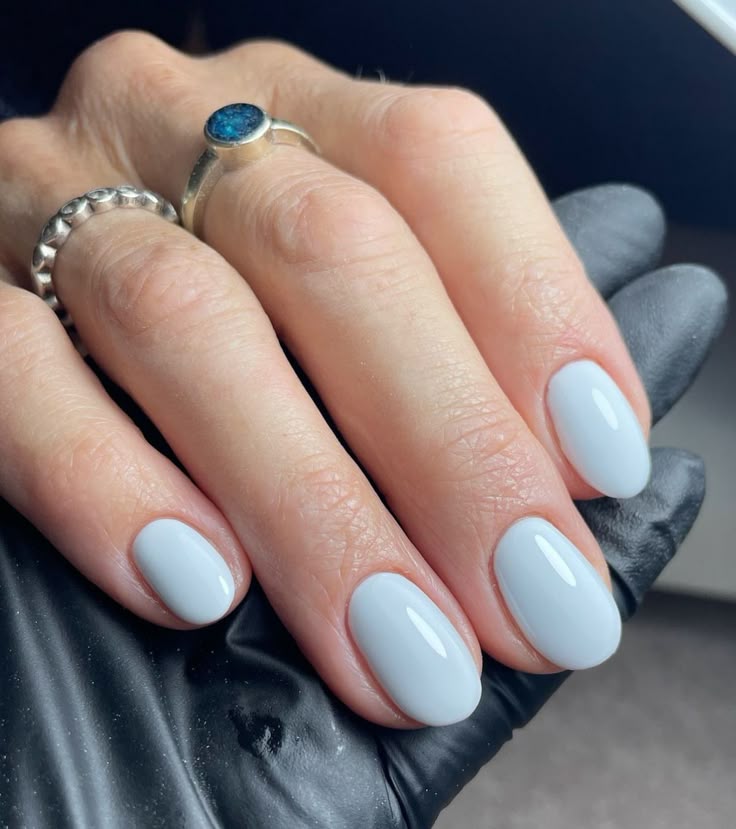 Mano con anillos y guante negro de fondo, mostrando uñas de longitud media con forma ovalada y esmalte en un tono blanco lechoso o gris muy tenue.