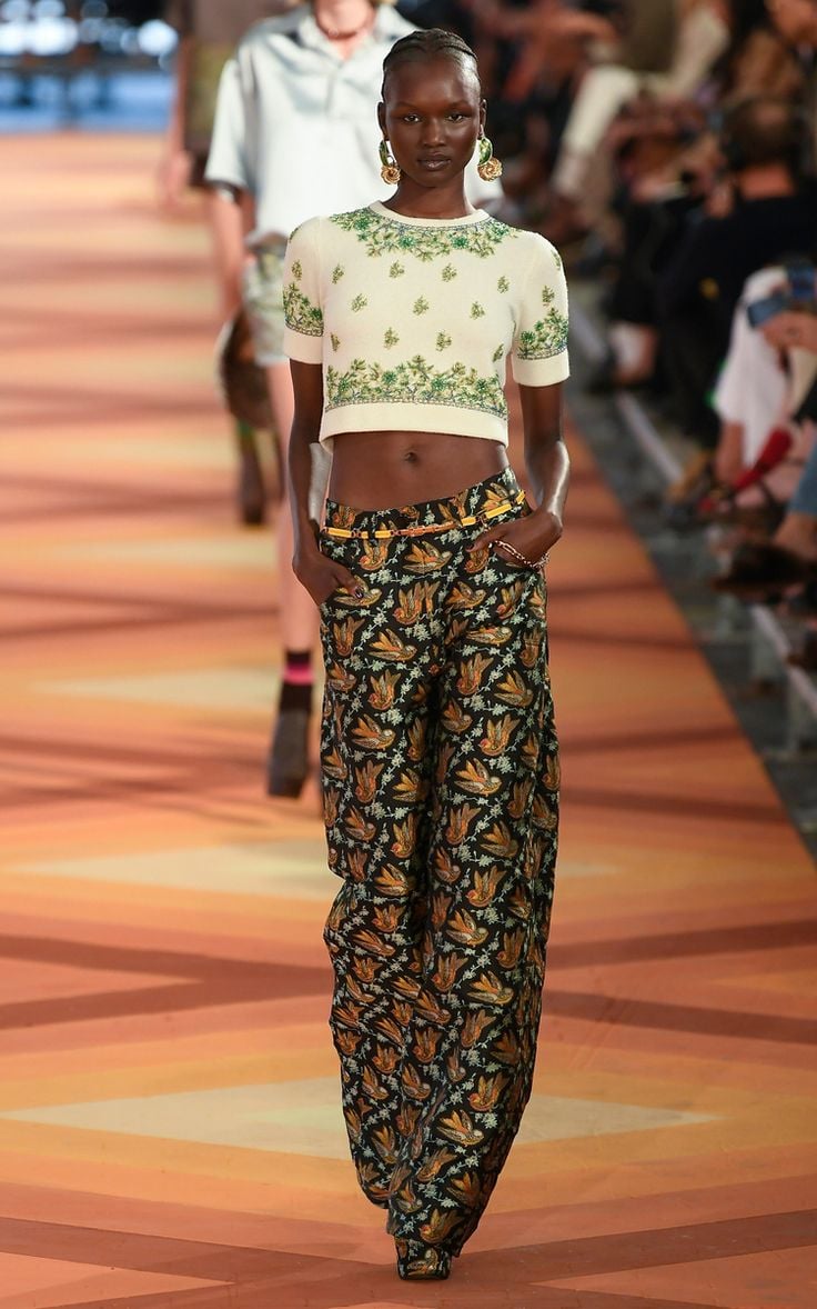 Modelo desfilando con un top corto de punto blanco con detalles bordados en verde y pantalones largos anchos con estampado de aves.