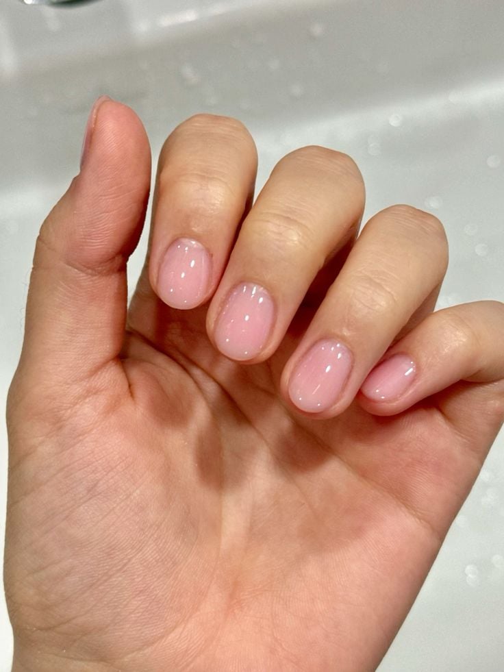 Mano mostrando uñas cortas y redondeadas con un esmalte rosa translúcido de aspecto natural y ultrabrillante.
