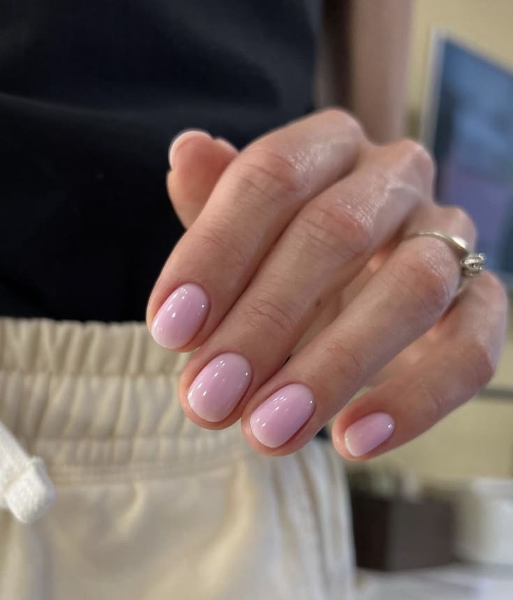 Mano con uñas cortas y pulidas con un esmalte en tono rosa pastel lechoso y brillante, usando un anillo fino de plata.